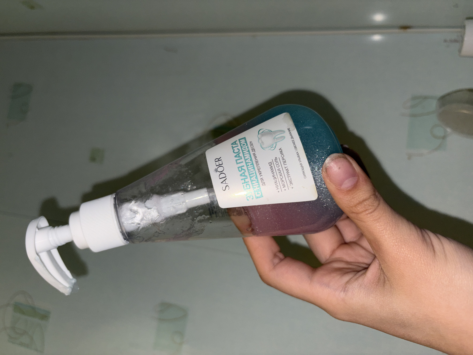 A hand holds a near-empty bottle of "SADOER" brand toothpaste, specifically identified as "ЗУБНАЯ ПАСТА СНИАЦИНАМИДОМ" which contains niacinamide for strengthening gums. The bottle is clear, allowing the remaining purple and blue toothpaste to be visible, and features a white pump dispenser. The background shows a flat, light green surface, likely a countertop or table. The location in the city of Прикамский, Russia is not identifiable from the image.