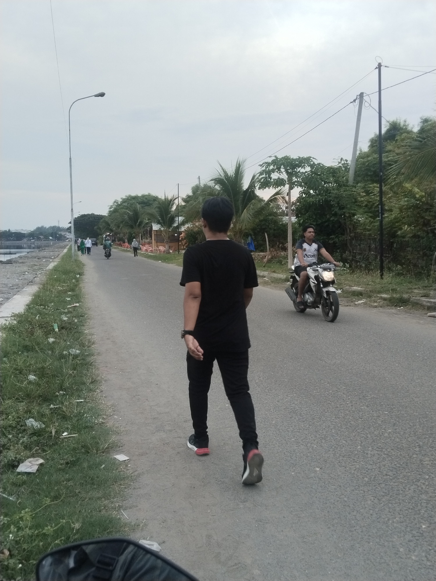 The image depicts a coastal road in Lhokseumawe, Indonesia, likely near the Ujong Blang beach area, given the proximity to the sea and palm trees. The scene is captured at an overcast time of day, possibly late afternoon or early morning. A person, seen from behind, is walking on the right side of the road, wearing a black t-shirt and black pants with red-accented sneakers. Further down the road, several other individuals are walking on the left side, dressed in casual attire, some wearing headscarves. A motorcycle with a rider wearing a white and black patterned t-shirt is traveling on the road in the same direction as the main subject. The environment includes a grassy verge along the left side of the road, littered with some debris, and a body of water to the far left, suggesting it's a coastal area. Palm trees and other vegetation line the right side of the road, with some structures that appear to be small shops or cafes visible in the distance. A street lamp is present on the left side of the road, and utility poles with overhead wires are visible in the background. The overall impression is a tranquil, everyday scene along a seaside thoroughfare.