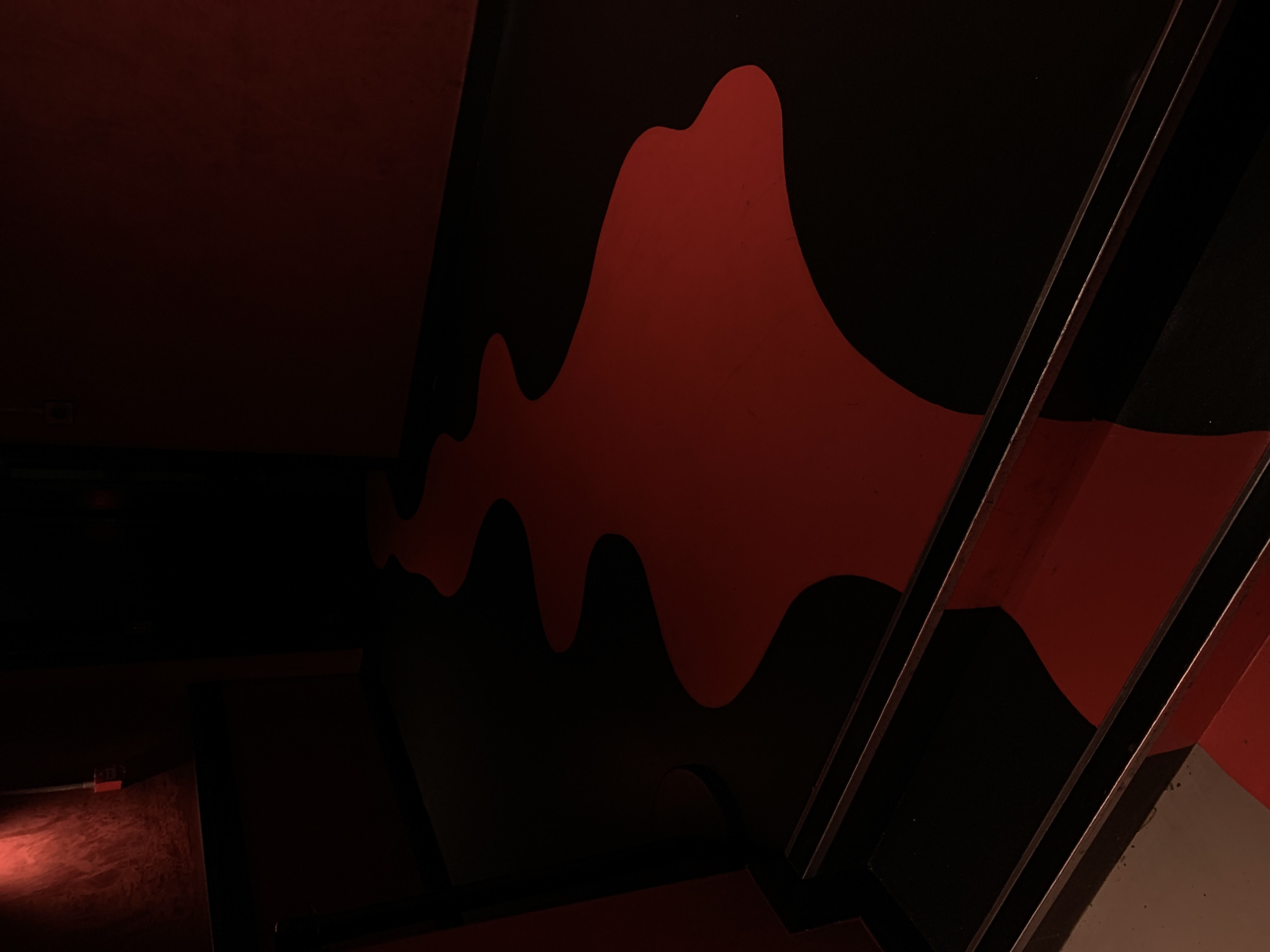 This image depicts a dimly lit, interior architectural space characterized by bold contrasts of black and deep red. The central feature is a large, abstract, wave-like or organic pattern painted in vibrant red against a black wall, dominating the mid-right portion of the frame. This dynamic, flowing design creates a strong visual impact within the dark environment.
On the right side, several parallel metallic beams or bars, possibly forming a railing or structural support, angle upwards diagonally. These elements reflect the ambient light, adding a metallic gleam to the composition. The overall lighting is low, casting significant portions of the image into shadow, particularly the upper and lower left areas.
In the bottom-left corner, a textured surface, possibly a floor or another wall, is faintly illuminated by a reddish glow, suggesting an unseen light source. A small, dark rectangular object, possibly an electrical outlet or box, is visible on this surface. No people or specific activities are depicted. The scene conveys a moody and atmospheric ambiance, reminiscent of a modern club or entertainment venue, consistent with locations found in London, United Kingdom. No visible text is present.