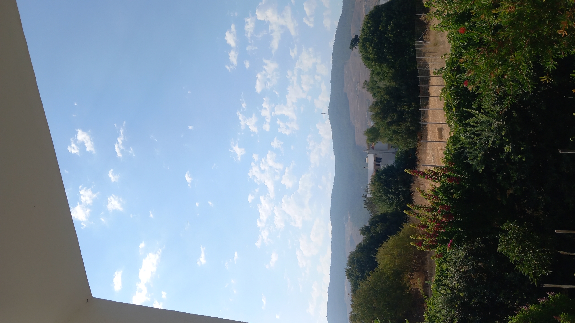 The image depicts a landscape scene with a clear blue sky filled with scattered white clouds. In the foreground, there are lush green bushes and trees, with some flowering plants displaying red blossoms. A fence is visible behind the vegetation, separating it from a dry, golden field. In the middle ground, a white building with multiple windows stands among more trees. The background features rolling hills covered in a mix of trees and sparse vegetation, with two tall communication towers on one of the hilltops. A white overhang is present in the upper left corner of the frame, suggesting the viewpoint is from under a structure, possibly a balcony or roof. The overall scene suggests an outdoor environment in a rural or semi-rural area, under a bright, sunny sky. The location is specified as Άγιος Νεκτάριος, Greece.