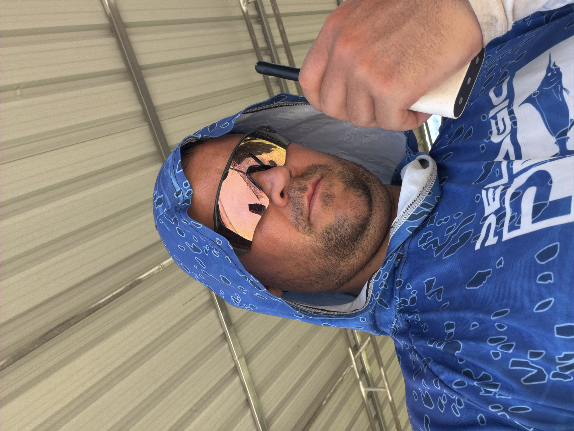 A close-up, low-angle shot shows a man wearing a blue hooded shirt with a white and blue pattern, holding a white cylindrical object in his right hand and a black object in his left hand. He is also wearing mirrored sunglasses. The background consists of a corrugated metal roof supported by metal beams. The man's face is partially obscured by his hood and sunglasses. He appears to be outdoors, possibly in a covered outdoor area. The lighting suggests it is daytime. The scene takes place in Baan Bo Phut, Thailand.
