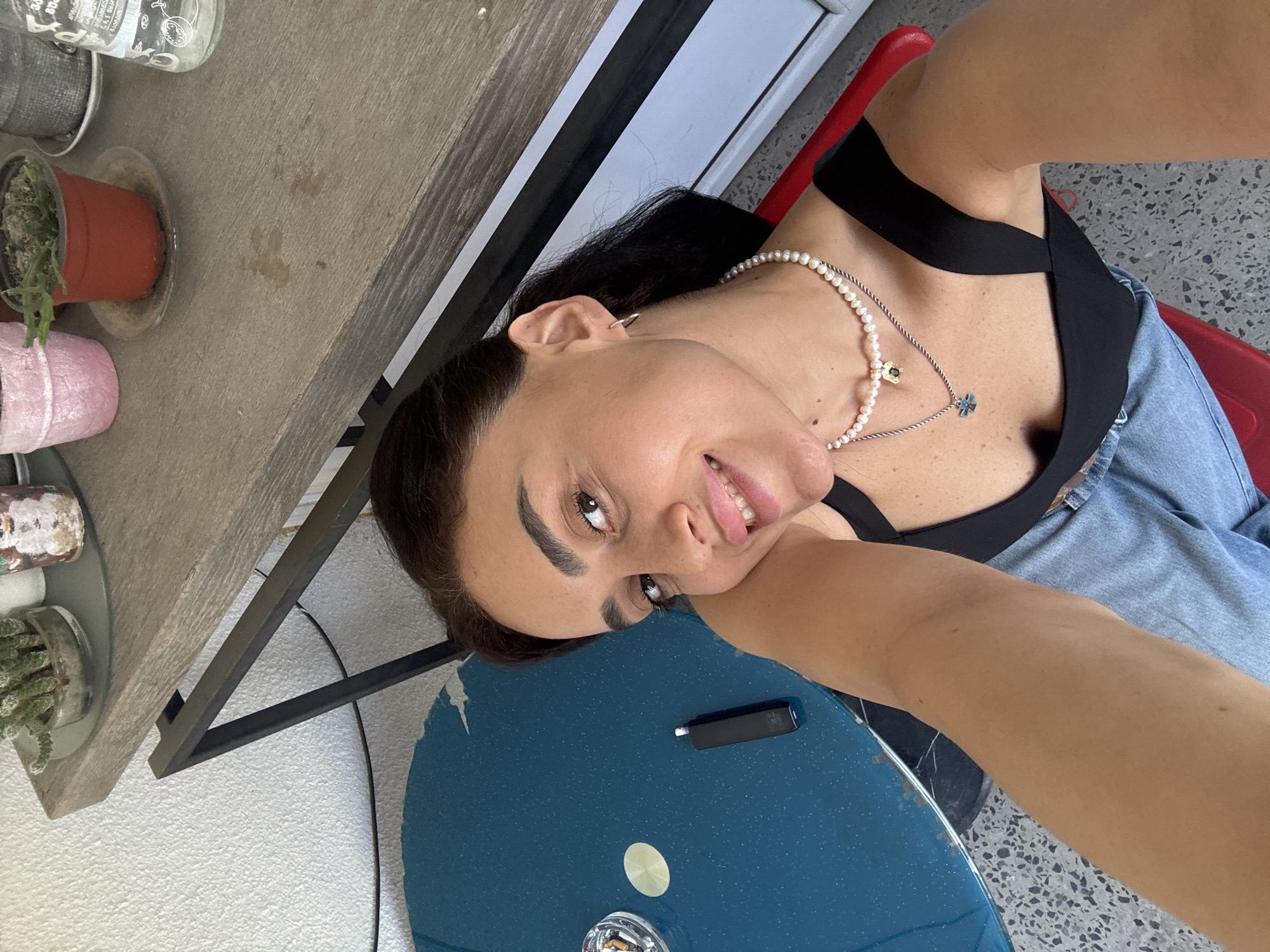A woman is taking a selfie in a relaxed, outdoor setting. She is wearing a black top and denim shorts, with a pearl necklace around her neck. Her hair is dark and pulled back. She has a friendly smile and appears to be enjoying herself. The background shows a table with some potted plants and a blue circular object, possibly a table or tray, with a small black device on it. The overall impression is casual and summery. The scene appears to be well-lit, suggesting daytime, and the concrete-like floor with speckles hints at an outdoor patio or balcony. There is no visible text in the image.