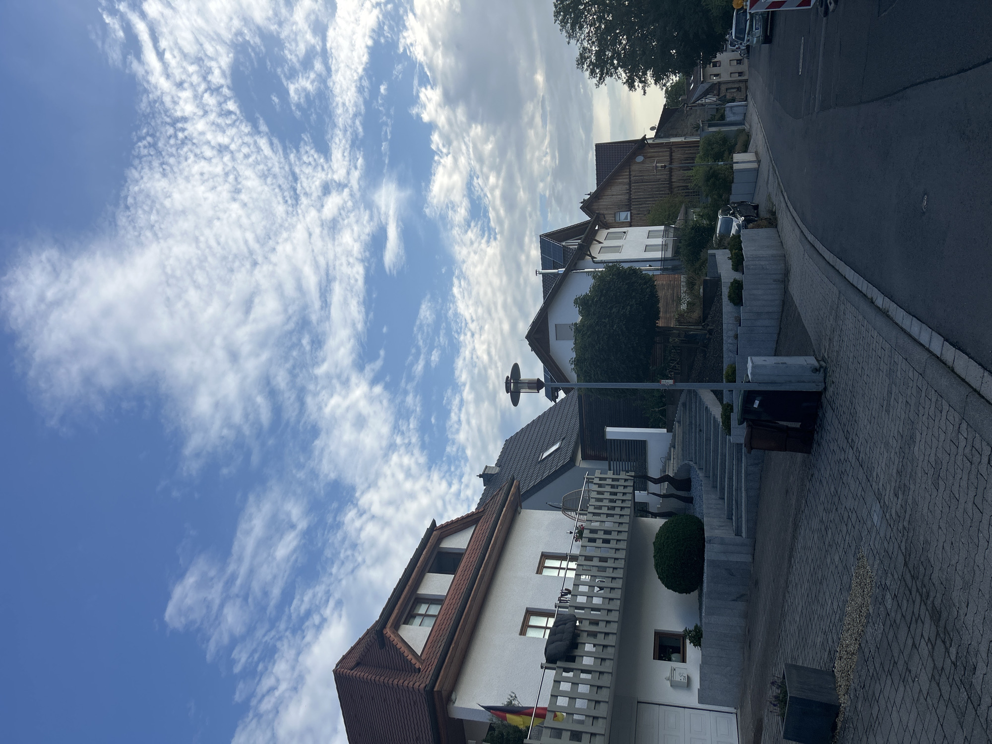 The image depicts a residential street in what appears to be a suburban or rural area, given the presence of houses, trees, and a clear sky. The architecture suggests a European setting, possibly Germany due to the flag visible on the first house. The street curves to the right in the distance, with houses lining both sides. The sky is blue with scattered white clouds, indicating fair weather. The foreground shows a paved walkway and a street. A white house with a red-tiled roof is prominent on the left, featuring a balcony with a railing. Several other houses of varying architectural styles and materials are visible further down the street. Some cars are parked along the road. The overall scene is one of a quiet, everyday setting.