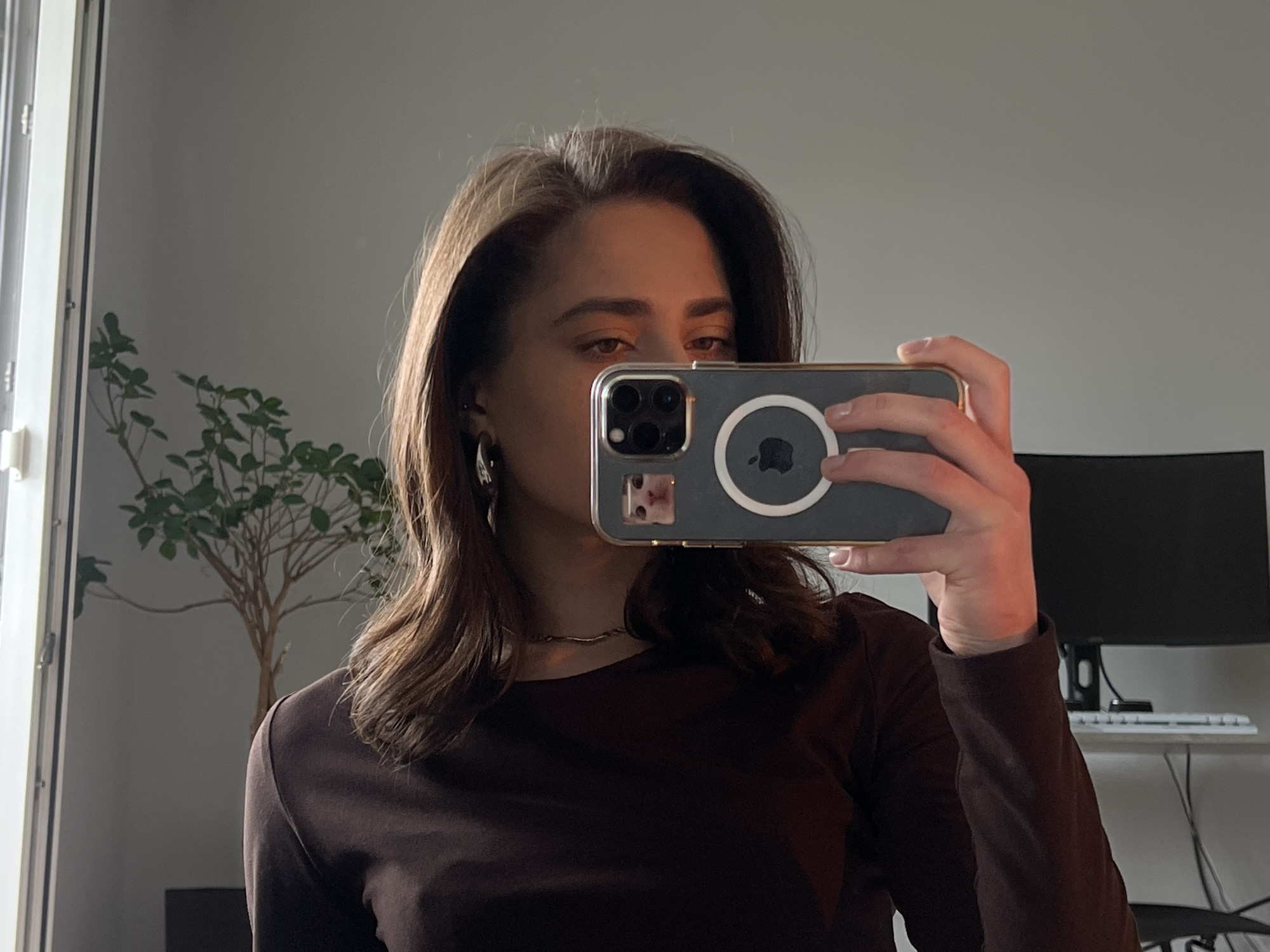 In the city of Ternopil, Ukraine, a young woman is taking a selfie in a mirror. She is holding up a smartphone with a magnetic accessory attached to the back, and is wearing a dark brown, long-sleeved shirt and hoop earrings. She has brown hair, which is styled in loose waves, and is looking directly at her phone's screen. The background includes a large window with a plant and a computer monitor on a shelf. The lighting suggests it is daytime, with natural light filtering in from the window.