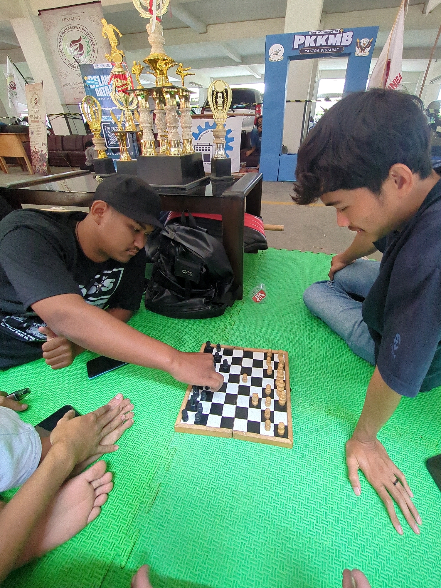 This image depicts an indoor scene, possibly a community event or sports competition, with elements suggesting it's located in Surakarta, Indonesia. In the foreground, two young men are seated on a bright green, textured mat, engaged in a game of chess. The man on the left, wearing a black t-shirt and a black baseball cap, is actively moving a chess piece on the board with his right hand. He is also holding a dark-colored smartphone with his left hand, positioned near the chessboard.
The man on the right, wearing a navy blue t-shirt and blue jeans, is looking intently at the chessboard. His right hand is resting on the mat, and his left arm is bent at the elbow. In the background, a table with a dark surface is visible, with several trophies displayed prominently on it. These trophies are golden and appear to be of various sizes and designs, indicating a competitive event.
Behind the trophies, a blue archway with the text "PKKMB" and "ASTRA VISTARA" is visible, along with banners featuring text in Indonesian, likely related to the event or organization. The overall environment suggests a public gathering space, potentially a hall or a covered area, with concrete flooring visible beyond the green mat. The presence of the trophies and banners strongly suggests an academic or organizational event.