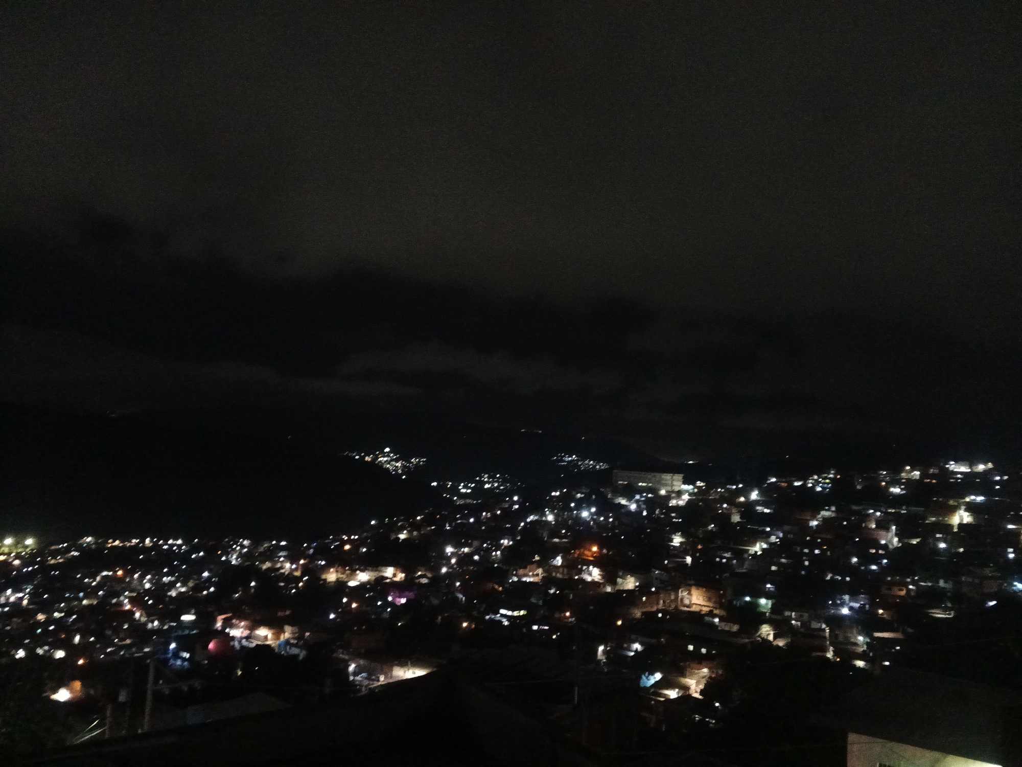 A night view shows an urban landscape in Caracas, Venezuela. The foreground is obscured by shadow, likely a building or hillside from which the photograph was taken. The middle ground reveals a dense collection of lights from houses, streets, and businesses. Above the city, the night sky is a dark gray mass, possibly a mix of clouds and light pollution. No people are visible in the photograph. The overall atmosphere is one of a sprawling city at rest, punctuated by the lights that mark its existence.