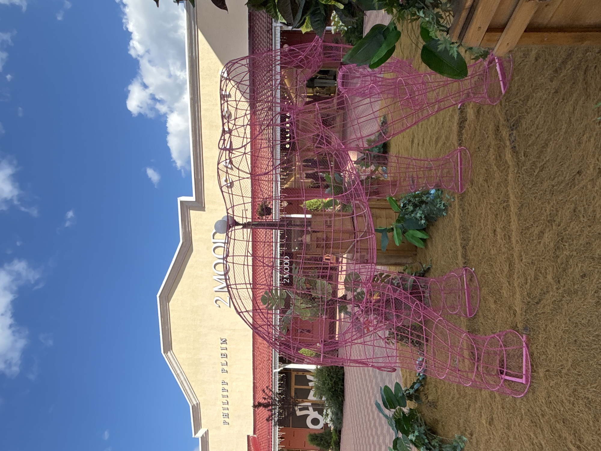 The image depicts an outdoor scene in front of a building with a beige facade and a red tiled roof. The building has signage that reads "PHILIPP PLEIN" and "2 MOOD". In the foreground, a large, pink wireframe sculpture of an elephant stands on a ground covered with dry, brown fibrous material. The elephant sculpture is hollow and its structure is made of interconnected wire loops and straight pieces. Green foliage and plants are visible around the sculpture and to the sides of the image. The sky above is blue with scattered white clouds. The scene appears to be a commercial area, possibly a shopping outlet or a street in a city. The provided context specifies the location as Zakharovo, Russia.