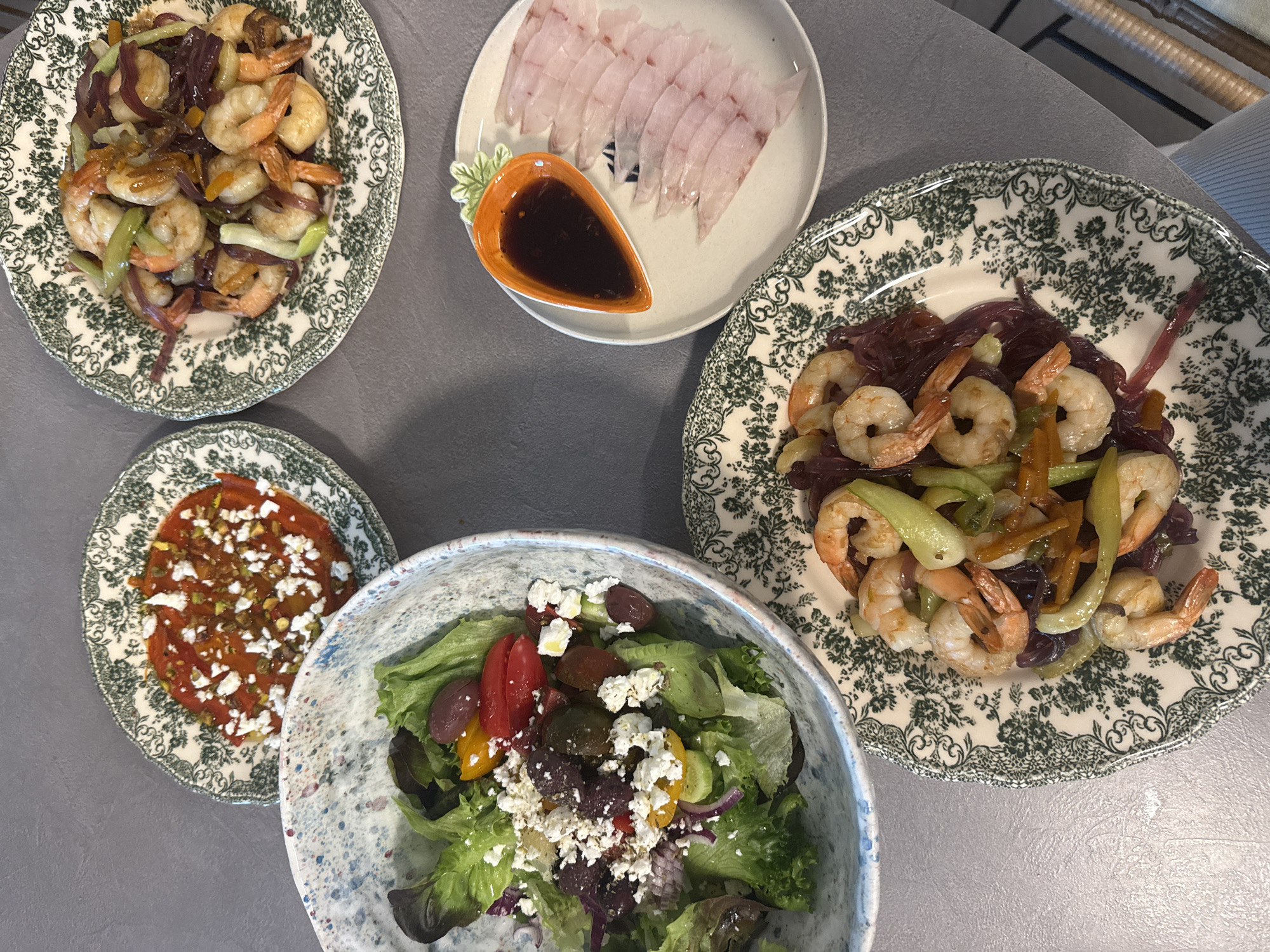 The image depicts a table with several plates of food, likely taken in Ramat Gan, Israel. There are four plates in the foreground, and a fifth visible in the background to the right.
The foreground plates are arranged in a rough square. Starting from the top left, there's a plate with shrimp and vegetables (possibly onions and celery) in a dark sauce. The next plate shows thinly sliced white fish, possibly sashimi, with a small dish of dark dipping sauce. To the right and slightly below is another plate with a shrimp and vegetable dish. The bottom left plate contains a tomato-based dish with what appears to be feta cheese on top. The bottom center contains a salad with lettuce, olives, tomatoes, and feta cheese.
The table has a grey or neutral-toned surface. A portion of a chair or other furniture is visible in the upper right. There is nothing obviously identifiable in the image that places it within Ramat Gan specifically; that location is purely from the prompt.