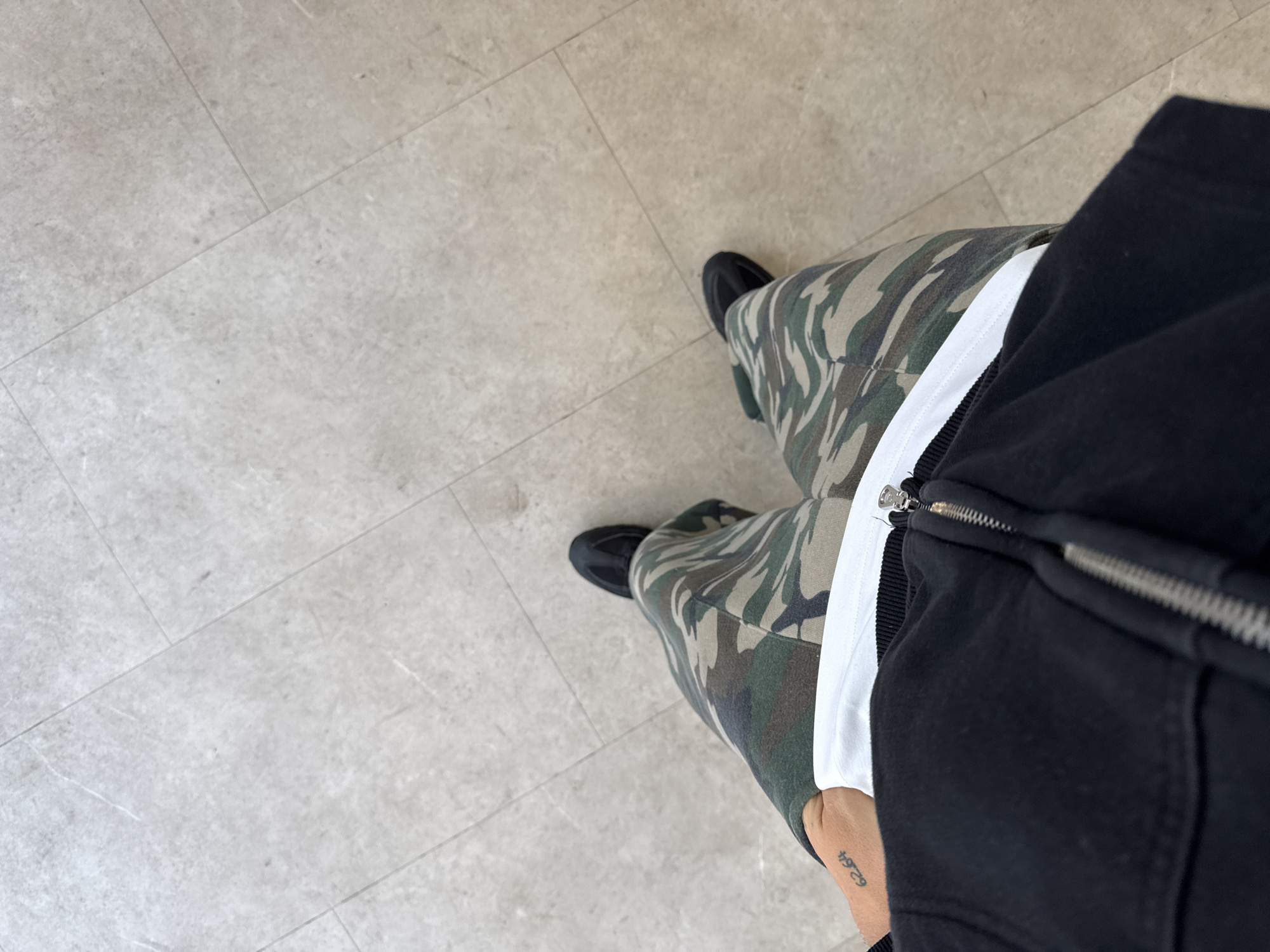 The image depicts a downward-angled view of a person standing on a light grey tiled floor. The individual is wearing camouflage trousers, black trainers, a white t-shirt, and a black zip-up hoodie. A tattoo is visible on the person's left wrist. The location is not identifiable as being in the city of London, United Kingdom. The scene includes a person standing on a tiled floor. The person's clothing is described. No specific actions or interactions are visible. The environment consists of a light grey tiled floor.