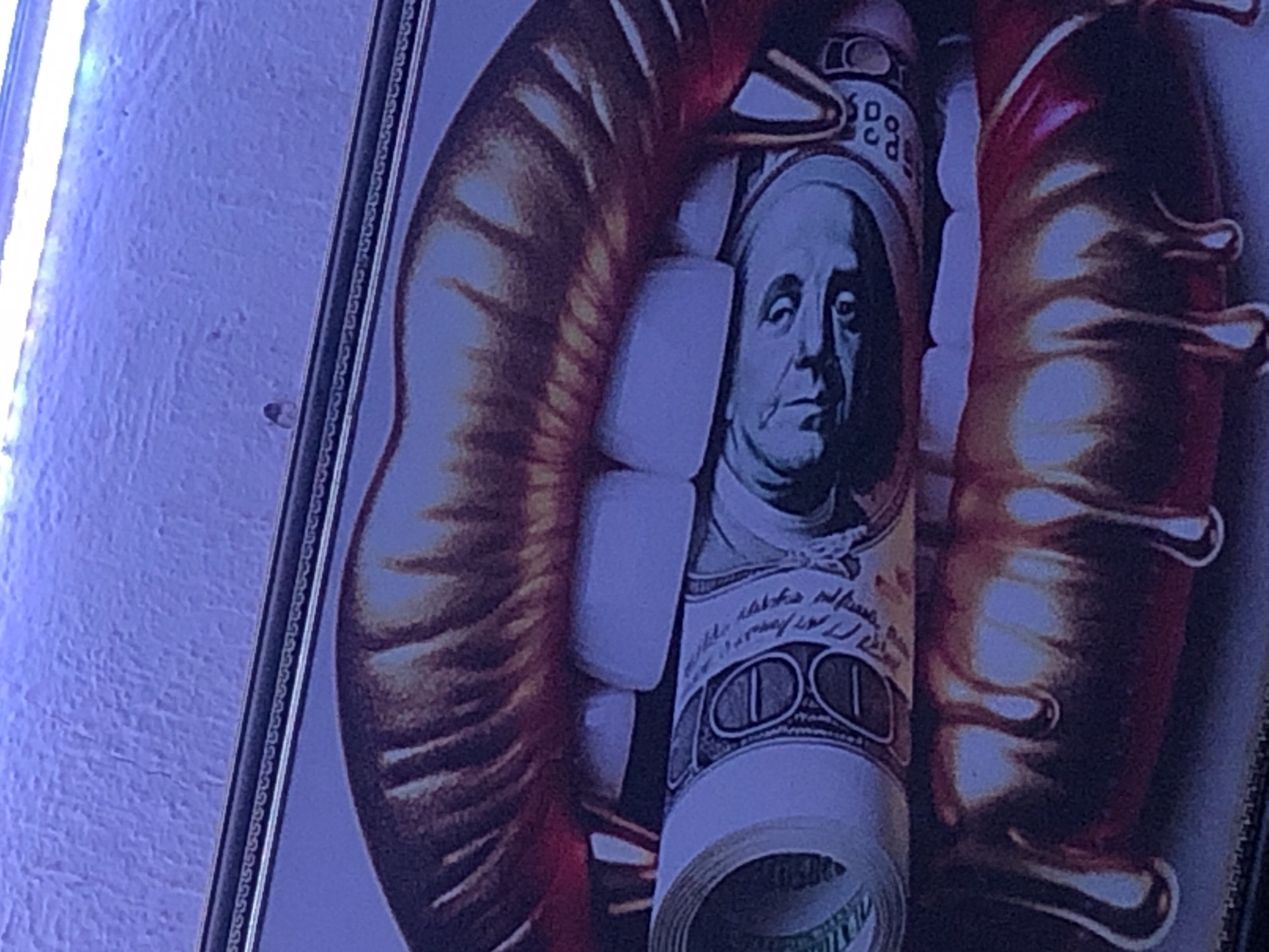 The image is a close-up of a framed artwork featuring a stylized depiction of human lips. The lips are depicted as glossy and dripping, rendered in shades of red and gold, suggesting a fluid or melting texture. A roll of US dollar bills, specifically $100 bills, is placed between the lips, with Benjamin Franklin's face clearly visible on one of the bills. The artwork appears to be illuminated by a cool, bluish light, casting a purple hue on the surrounding textured wall. There is no visible indication of a specific location in New Stadium, Nigeria, within the artwork itself or its immediate environment.