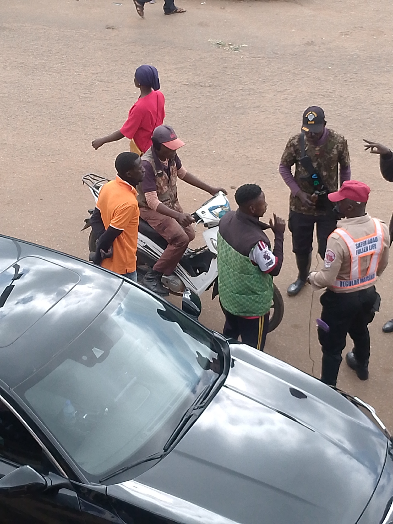 The image depicts an outdoor scene in Bukuru, Nigeria, likely a street or open area with a sandy, gravelly ground. Several individuals are present, along with a motorcycle and a car.
In the foreground, a dark-colored car is visible, with its windshield reflecting the sky and clouds. A portion of the car's hood shows a textured surface with a subtle shine.
A group of people are gathered in the middle ground. One person, wearing an orange shirt, stands with their back to the camera, arms held behind their back. Next to them, a person on a motorcycle, wearing a camouflaged vest and a red cap, is partially visible. Another individual, wearing a green checkered vest over a jacket and a blue headscarf, is speaking, gesturing with their right hand. To their right, a person in a beige uniform with an orange vest and a red cap is facing the gesturing individual. The vest has reflective stripes and text, including "SAFER ROAD FULLER LIFE" and "REGULAR MARSHA". This person is also wearing black trousers and black shoes.
Further back, another person in a camouflaged shirt and a dark cap with a logo is standing. Behind them, a person in a bright pink shirt and a dark headscarf is walking away from the camera. Some blurry figures are visible in the background, suggesting more activity or people present. The environment appears to be an unpaved area, possibly a market or a roadside.