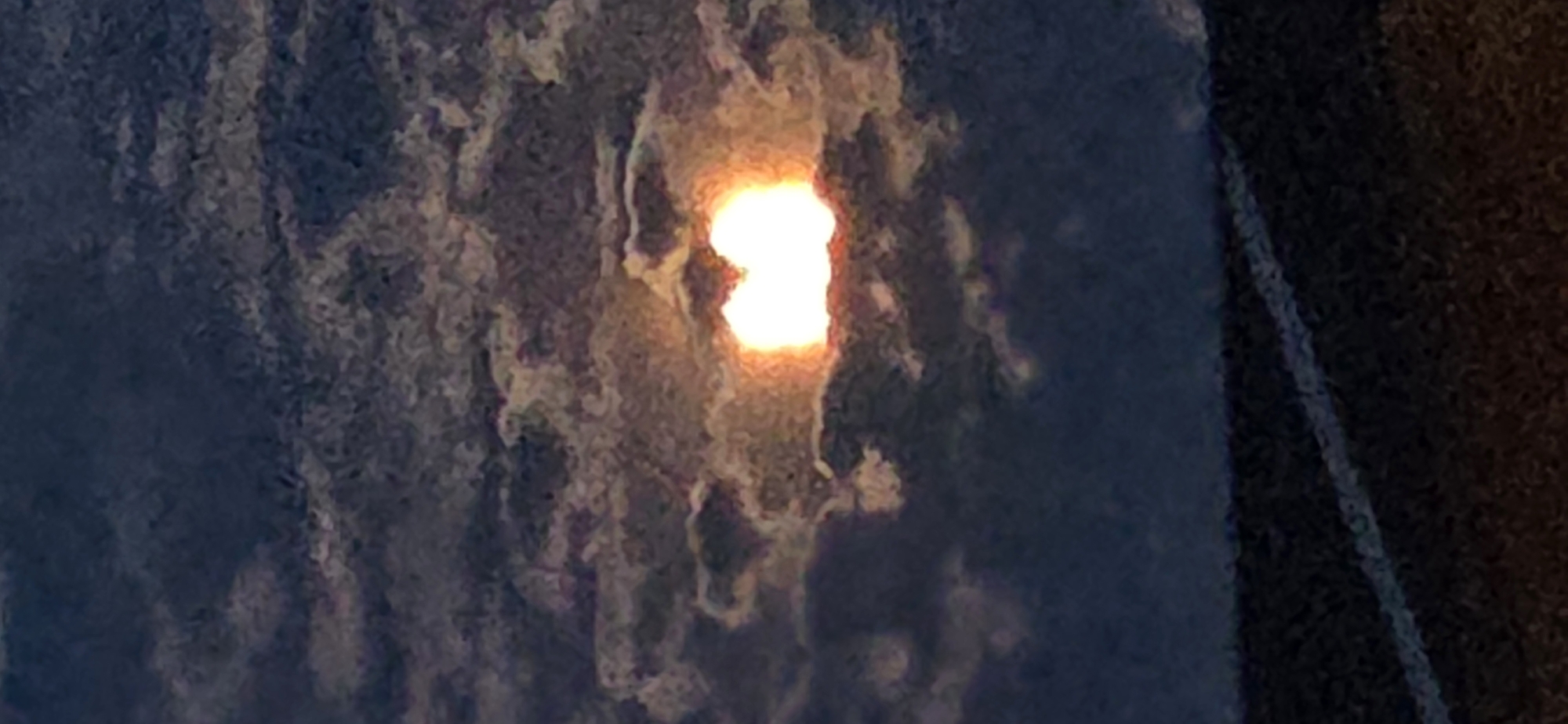 The image depicts a sky with thick, dark clouds partially obscuring a bright, yellow-orange celestial body, possibly the sun or moon, which appears as a radiant core surrounded by lighter clouds. The celestial body is centrally located in the upper half of the image and has a somewhat diffuse, glowing appearance, with hints of red and pink at its edges. The clouds are textured and layered, creating a sense of depth and drama in the sky.
In the lower portion of the image, a dark surface with a diagonal white line is visible. This surface appears to be a road or pathway, with the white line likely marking a lane or boundary. The texture of this surface is grainy and dark, suggesting it might be asphalt or a similar material, possibly illuminated by ambient light from the sky or an unseen source. The overall lighting suggests either dusk or dawn, or perhaps a heavily overcast day with the sun or moon shining through.
The image does not contain any people, animals, buildings, or specific landmarks that would allow for precise localization within Eeklo, Belgium. The scene is primarily focused on the atmospheric conditions and a distant terrestrial element.