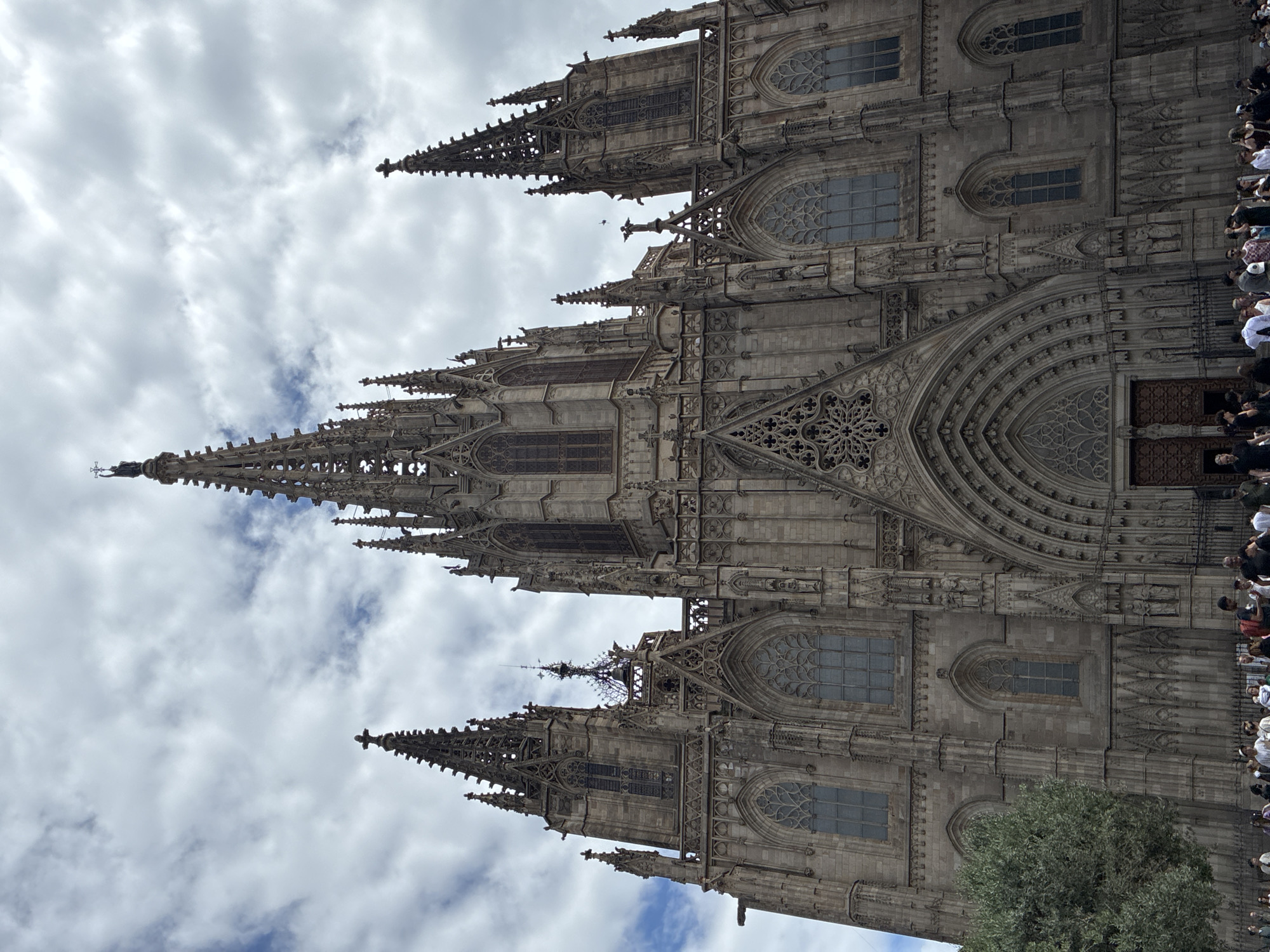 The image shows the Barcelona Cathedral, a Gothic cathedral in Barcelona, Spain. The cathedral is made of gray stone and has a tall, pointed spire. There are people in the foreground, near the entrance, suggesting the cathedral is open to the public. The sky is cloudy. A tree is partially visible in the bottom right corner.