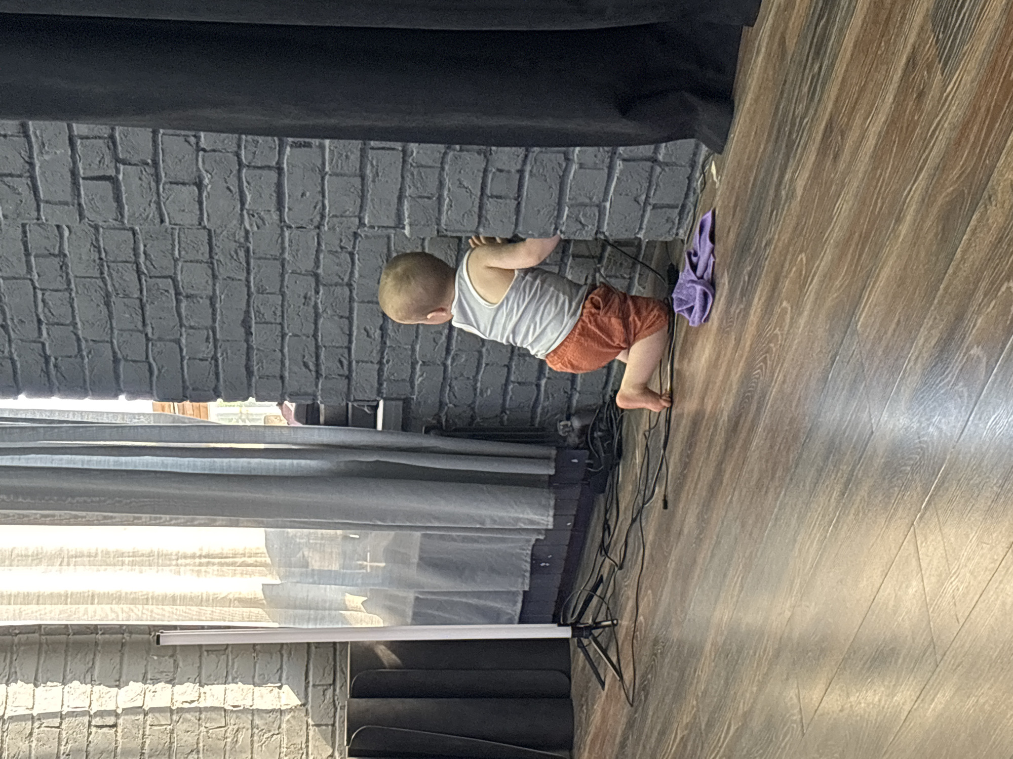 The image shows a toddler in a grey tank top and orange shorts leaning against a grey brick wall. The toddler is on a wooden floor. To the right of the toddler, a purple cloth is draped over a black cable. Behind the toddler, sheer white curtains are visible, with sunlight filtering through them, suggesting it is daytime. The overall scene appears to be indoors, possibly in a studio or a room with a brick wall feature and wooden flooring. The toddler's posture suggests they are actively exploring or playing. No text is visible in the image. The location context is Naberezhnye Chelny, Russia.