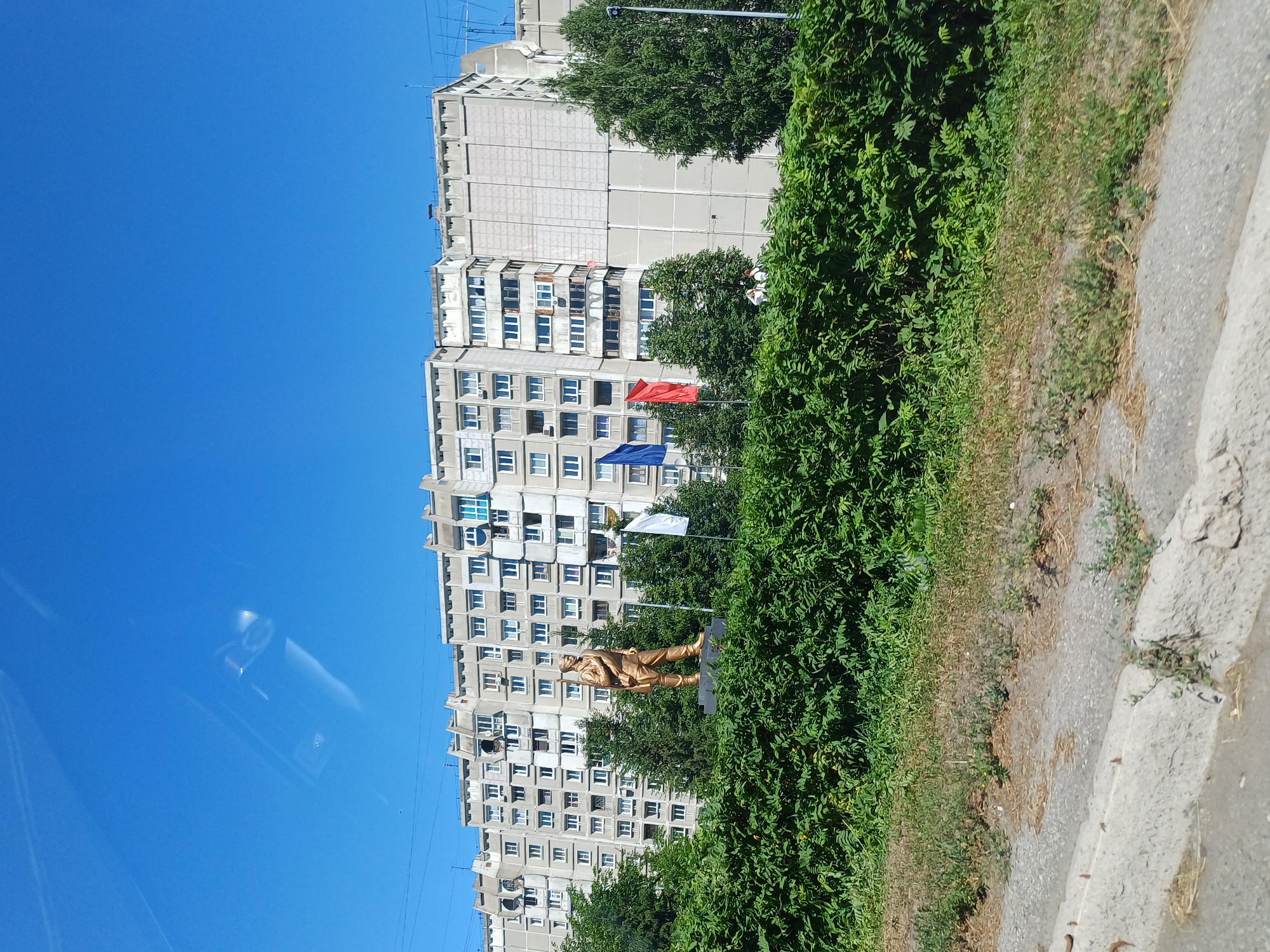 A golden statue of a soldier stands in front of a long, multi-story apartment building with a light-colored facade and numerous windows. The building appears to be of Soviet-era architecture. Green trees and bushes are visible in front of and to the sides of the building, partially obscuring the lower floors. Several flags are planted near the statue, including blue, red, and white ones. The sky above is a clear, bright blue. The foreground shows a concrete barrier and a patch of grass. The image is taken from a slightly elevated perspective, possibly from a vehicle.