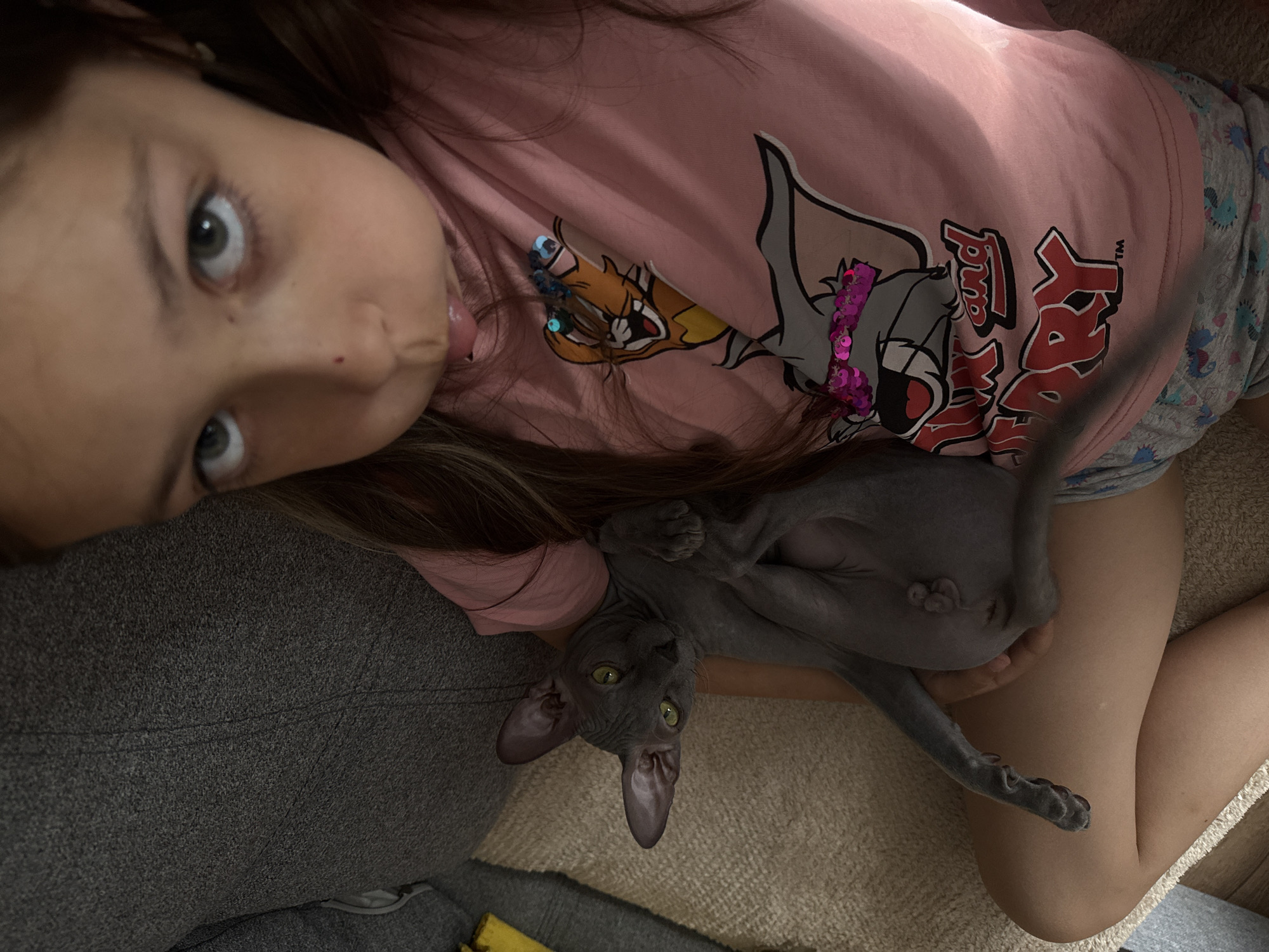 A young girl lies on a carpet, holding a grey hairless cat. The girl is wearing a pink shirt with a Tom and Jerry design, and grey patterned shorts. The cat is lying on its back, with its tail extending across the girl's leg. The background includes a grey sofa and a section of carpet. There is no evidence to confirm the location is Votkinsk, Russia.