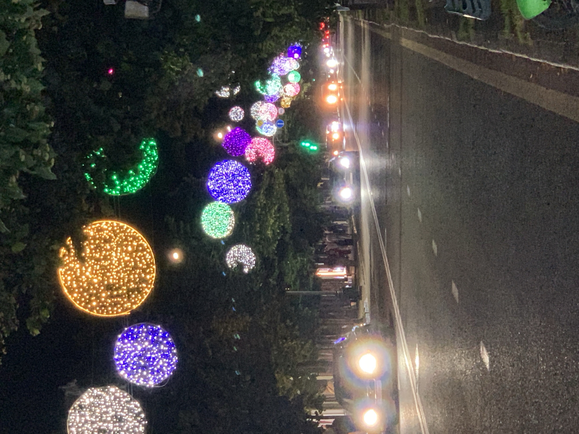 An eye-level, long shot captures a street at night, illuminated by a festive display of colorful, spherical lights strung across trees lining both sides of the road. The lights are predominantly white, purple, orange, and green, creating a vibrant, almost magical atmosphere. The road surface is wet and reflects the light from the overhead decorations and the headlights of passing cars, suggesting recent rain. Several vehicles are visible on the road, their headlights casting bright beams forward. On the sidewalks, figures can be seen gathered, their forms indistinct in the low light. The overall scene depicts a lively urban environment enhanced by decorative lighting.