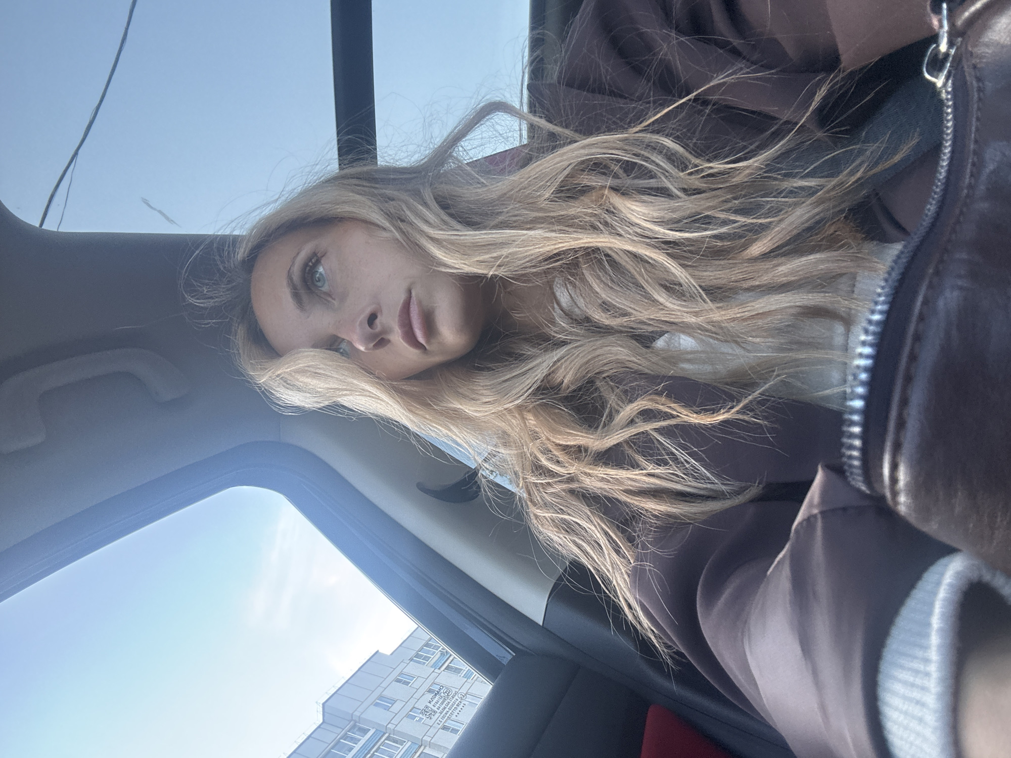 An overhead interior shot captures a young woman with blonde hair looking upwards from inside a vehicle. The woman is wearing a brown jacket and a light-colored top. The vehicle's sunroof is open, revealing a clear blue sky with a few wispy clouds. In the background, a multi-story residential building is visible, suggesting an urban or suburban setting, consistent with the city of Chaikovsky, Russia. The scene appears to be captured during daylight.
