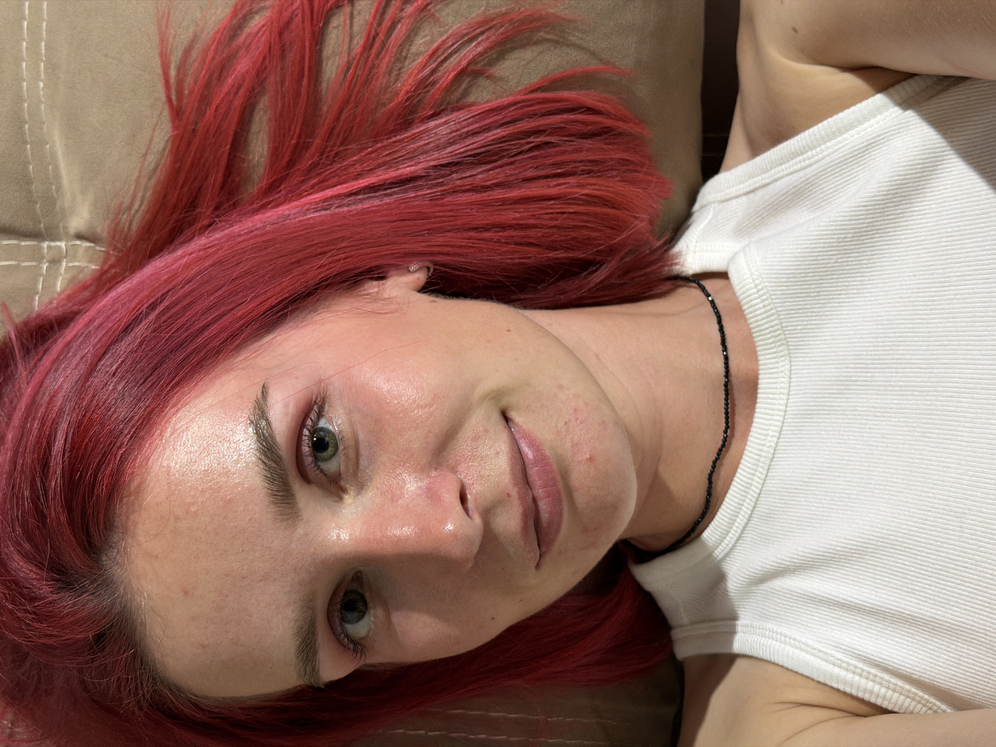 The image shows a close-up, overhead shot of a young woman with bright red hair lying down. She is wearing a white ribbed tank top and a delicate black beaded necklace. Her face is turned towards the camera, and she has a slight smile. Her skin appears fair with some redness and minor blemishes. Her eyes are open and looking directly at the viewer. The background is a textured, light brown surface, possibly a cushion or upholstery, with some visible stitching. The lighting suggests an indoor environment. No specific location in Chaikovsky, Russia can be determined from the image itself.