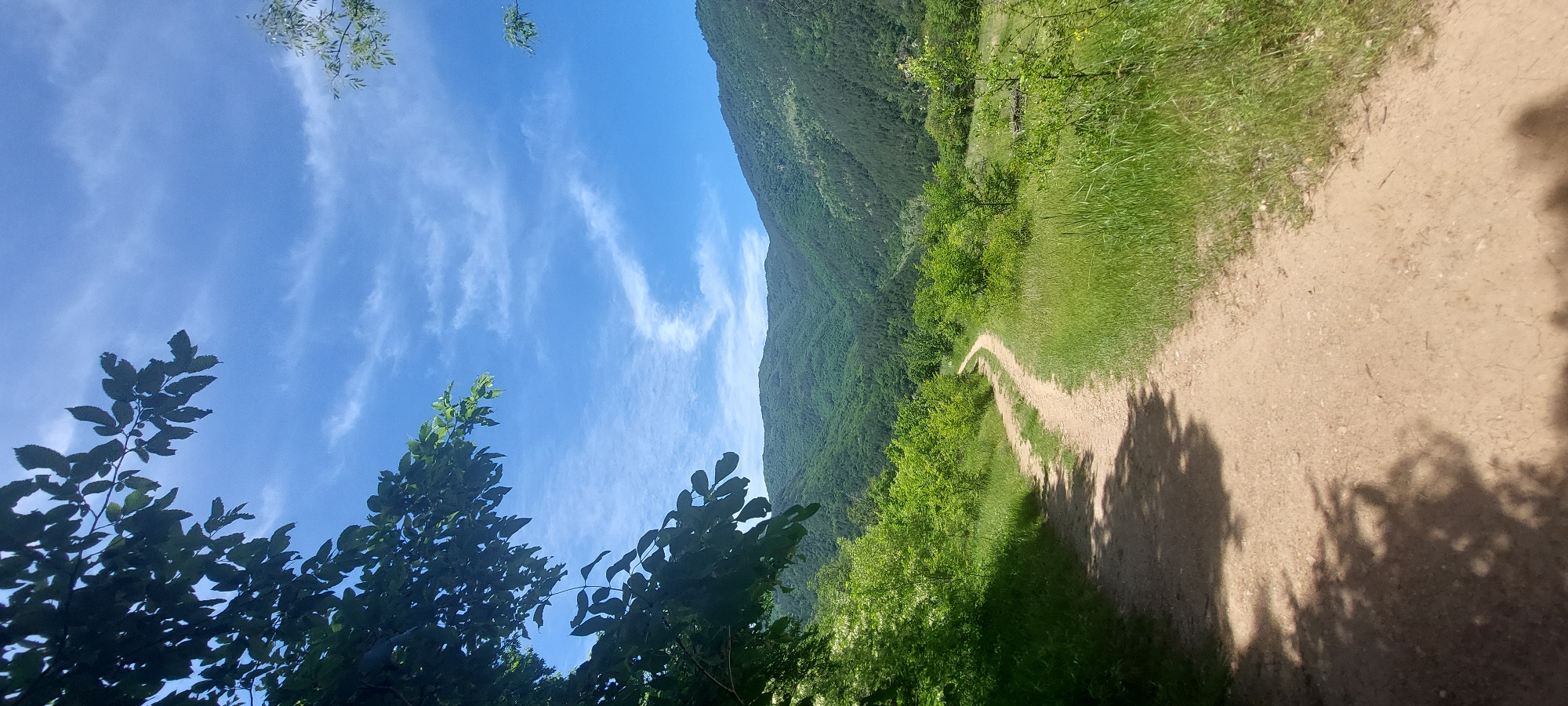 The image captures a dirt trail leading toward a green mountain or large hill, possibly in Bachkovo, Bulgaria, given the request context. The sky is blue with scattered wispy clouds. Trees and vegetation line both sides of the path. Shadows from the trees fall across the trail.