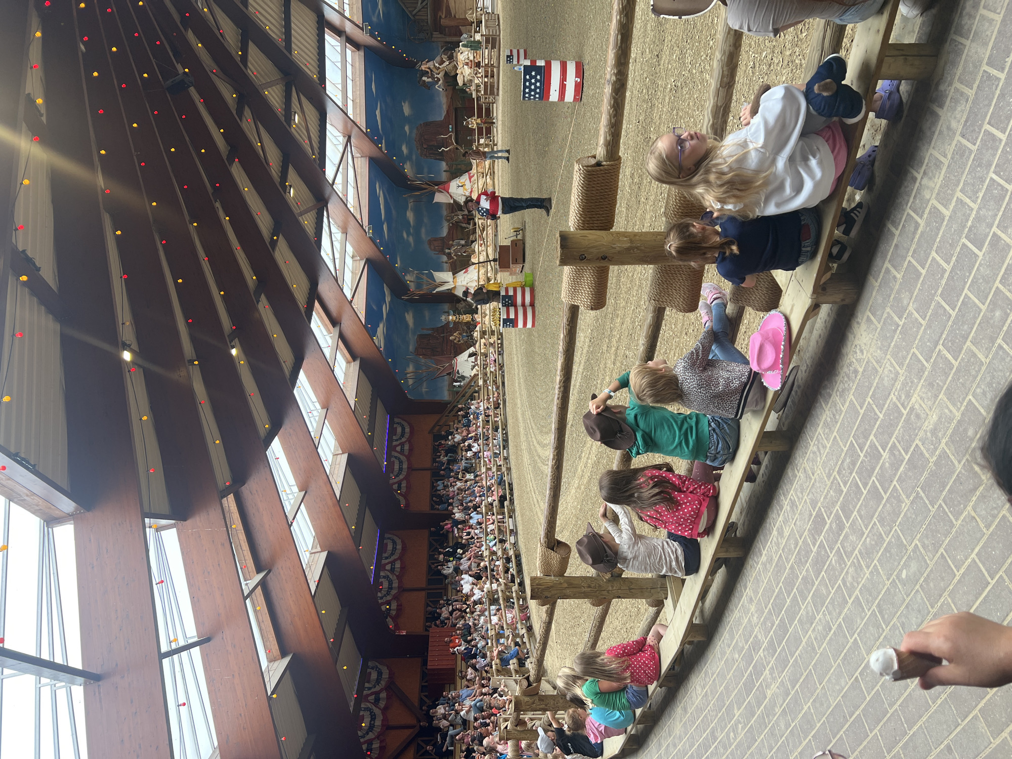 The image depicts an indoor rodeo arena in Collendoorn, Netherlands, hosting a performance for an audience. The arena is characterized by its sand-covered floor and a rustic wooden fence. A large crowd of spectators, including many children, are seated on tiered wooden benches along the perimeter of the arena, facing the performance area. Many of the children are wearing cowboy hats, suggesting a Western-themed event. In the arena, several performers are visible, including a man in a red, white, and blue shirt speaking into a microphone, and others dressed in Western attire. American flag-themed barrels are placed around the arena. The ceiling of the arena is high and features exposed wooden beams and rafters, with decorative red and yellow lights strung across it. The background of the arena is a large mural depicting a Wild West landscape with teepees, desert scenery, and figures on horseback. The overall atmosphere appears festive and engaging, with the performance drawing the attention of the seated audience.
