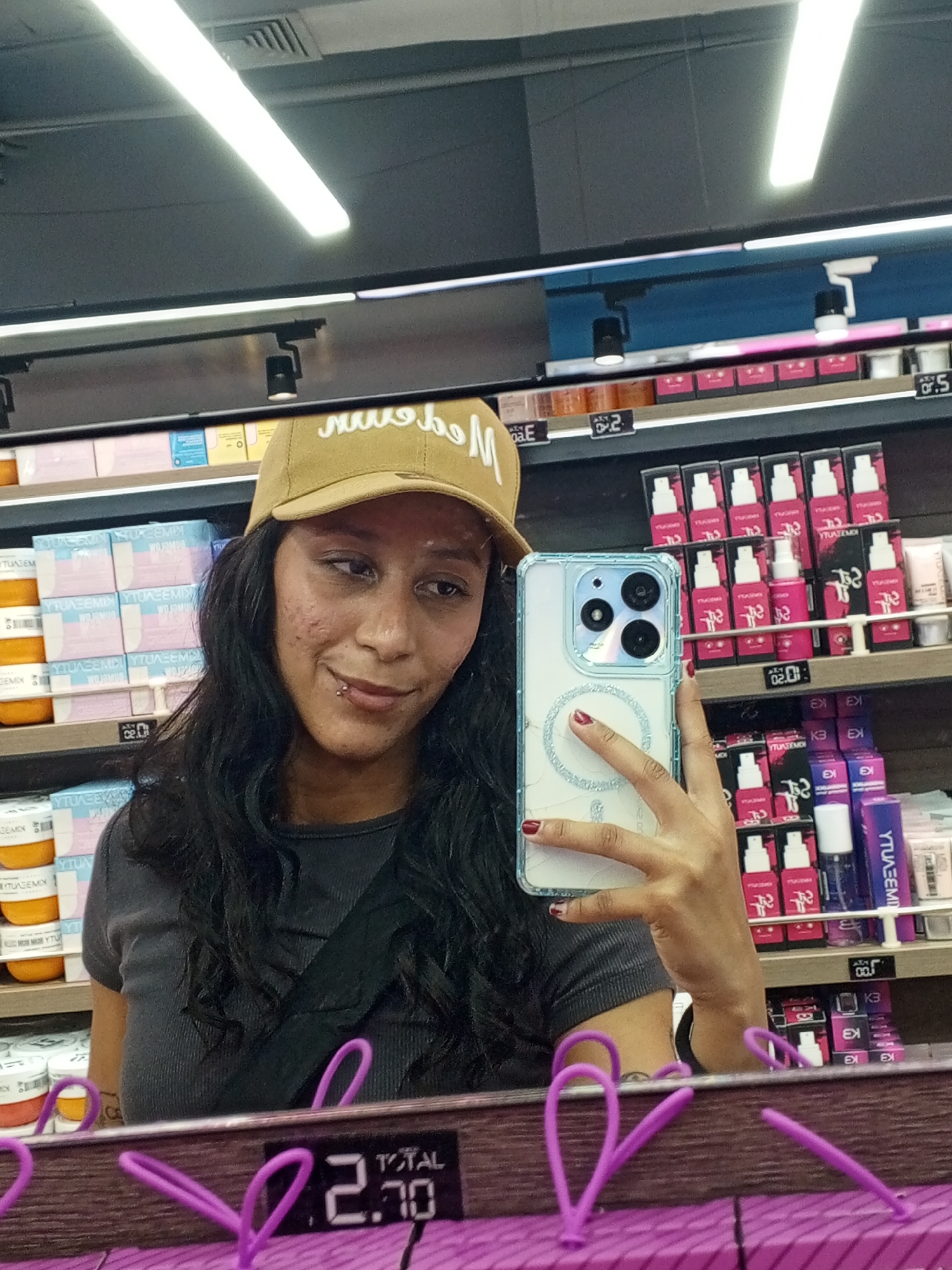 The image shows a person holding a smartphone, seemingly taking a selfie, in what appears to be a retail environment based on the shelved merchandise in the background.
The person is wearing a tan-colored baseball cap and a gray top. There's a black strap across their chest. The smartphone they are holding has a clear case with a ring accessory. The shelves behind them are stocked with various cosmetic or personal care products.
Reflected in the foreground is a price display showing "2.70," likely Venezuelan Bolívares, in addition to a portion of a purple display or stand. The products on the shelves behind the person appear to be organized by brand and type.
Since the image is a selfie taken within a retail setting, it's difficult to precisely determine the location to the city of Porlamar, Venezuela without additional information such as store signage, familiar brands or architectural details unique to the area.