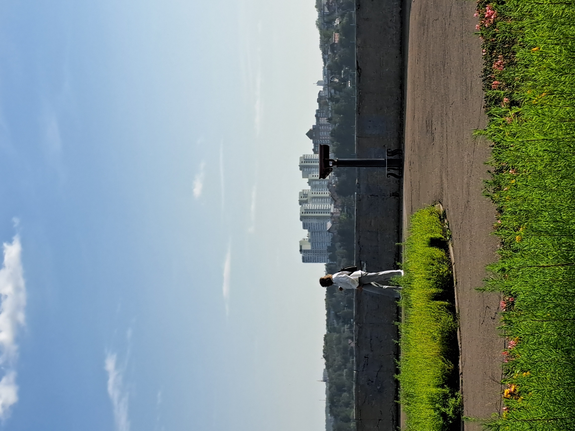 The image depicts a woman in white clothing and grey pants walking along a paved path next to a low concrete wall. Behind the wall, lush green bushes and trees form a dense line, leading to a cityscape in the distance. The cityscape features several multi-story buildings, including one particularly large, light-colored complex. A binocular viewer stands on a pedestal in front of the wall. The sky is blue with scattered white clouds. The foreground of the image is filled with a dense green plant life, with hints of pink and yellow flowers. This scene is likely located at the Angara River embankment in Irkutsk, Russia, a popular scenic spot for residents and tourists.
