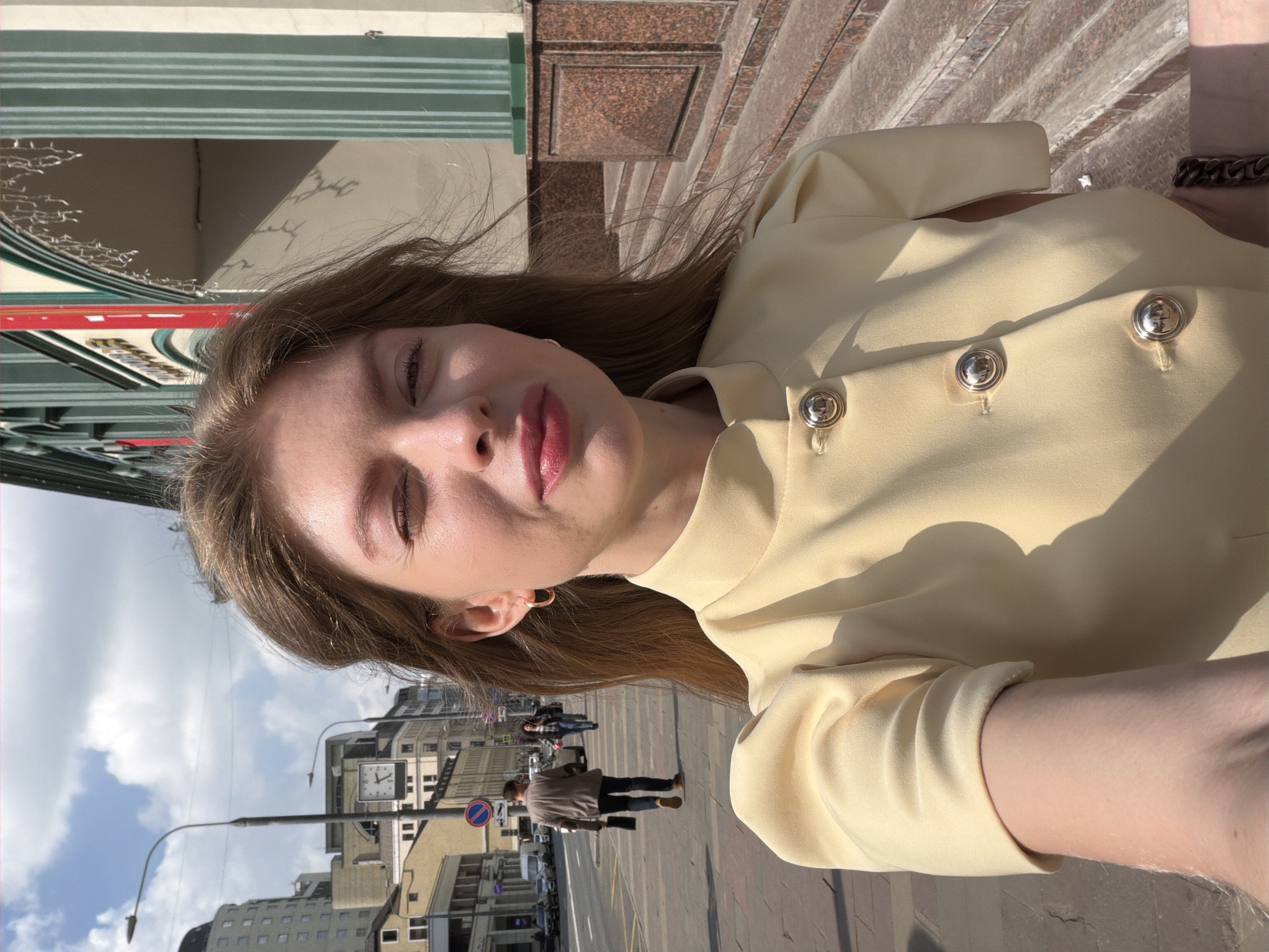 A woman takes a selfie in Moscow, Russia. She is wearing a light-yellow blouse with three large silver buttons. Her hair is brown and styled around her shoulders, and she is wearing hoop earrings. The background includes several buildings, one with a large clock face. Pedestrians are visible on the sidewalk. Street lamps and various street signs are also in view. The sky is blue with scattered clouds.