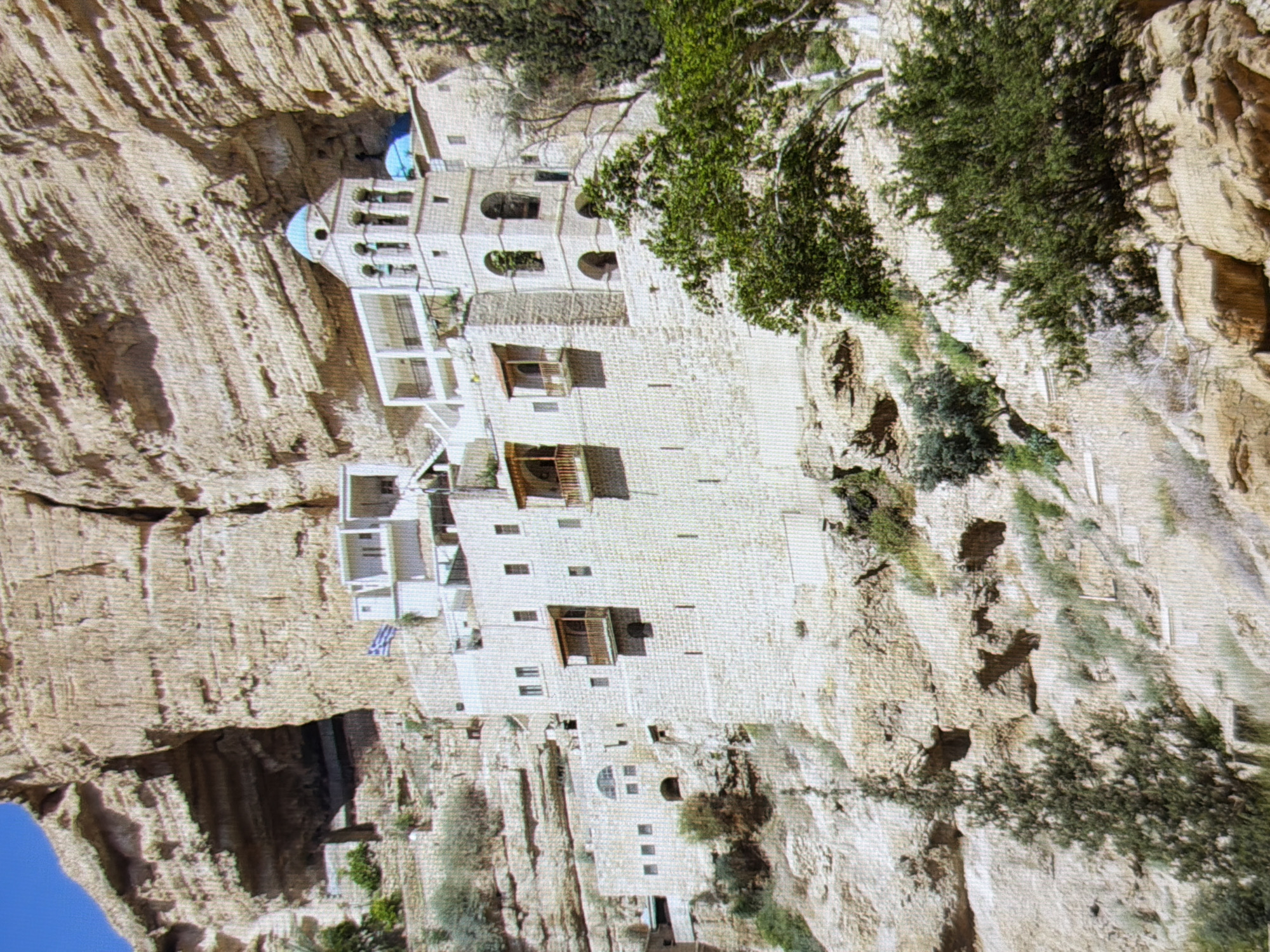 This image depicts a monastery built into a cliff face, likely in a rocky, arid region. The monastery is constructed of light-colored stone and features multiple levels with windows, balconies, and arched openings. One prominent section has blue domes, suggesting a chapel or religious structure. Green trees and vegetation are visible growing from the cliff and around the monastery, adding a touch of life to the stone facade. The lighting suggests it is daytime, with clear weather. A small blue and white flag is visible on one of the lower sections of the monastery. The overall scene conveys a sense of ancient architecture nestled within a natural, rugged landscape. There are no people visible in the image, and no text is discernable. The orientation of the image is rotated.