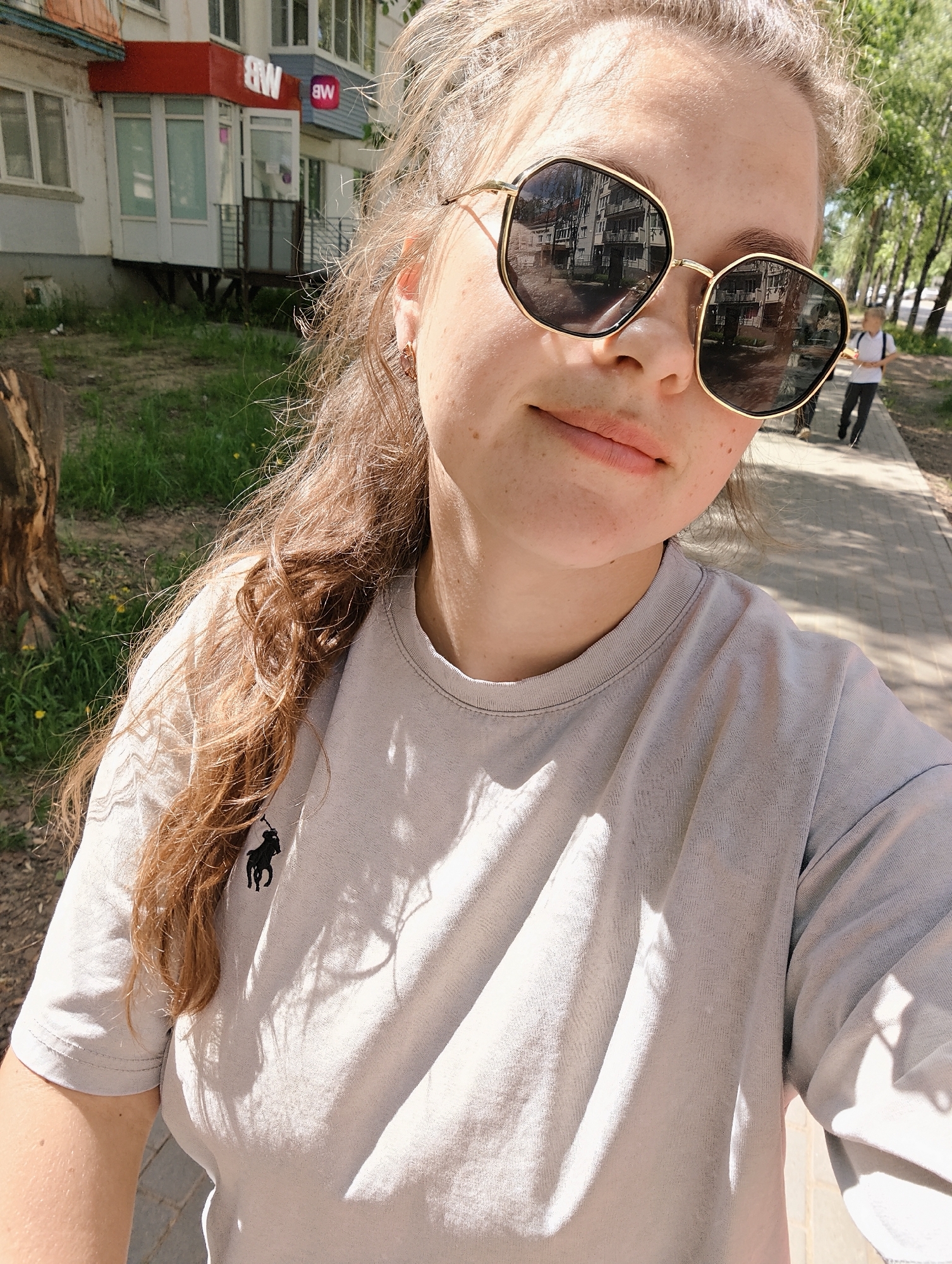 A young woman poses for a selfie outdoors on a sunny day. She is wearing large, geometric sunglasses that reflect a building with awnings and windows. She has light-colored hair partially tied back, and is wearing a pale grey t-shirt with a dark logo on the chest. The woman is standing on a brick sidewalk adjacent to a grassy area with trees. In the background, a person wearing a white shirt walks away from the camera on the sidewalk. The location is Chaikovsky, Russia; the reflection in her glasses shows a building with signage bearing the letters "BW" in Cyrillic.