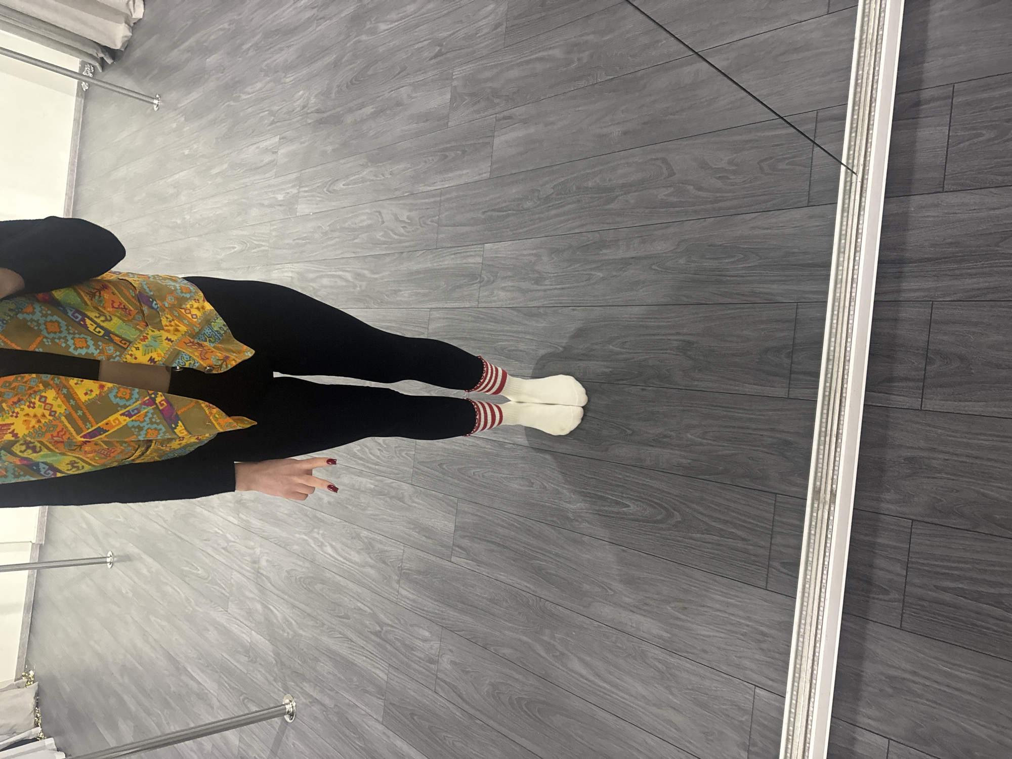 An individual is depicted standing in what appears to be an indoor pole fitness or dance studio. The floor consists of grey, wood-patterned laminate planks. Several chrome-plated vertical poles are visible, extending from the floor upwards. The person is photographed from the mid-torso down, wearing a black long-sleeved top, black leggings, and white ankle socks with two red horizontal stripes at the cuff. A patterned vest, featuring a multi-colored geometric or ethnic design in shades of orange, yellow, green, and blue, is worn over the top. The individual's left hand, with fingernails painted red, is visible at their side. The bottom edge of a large, framed mirror, which reflects the person and a portion of the floor, is prominent at the bottom of the image. The background reveals light-colored walls and partial white drapery.