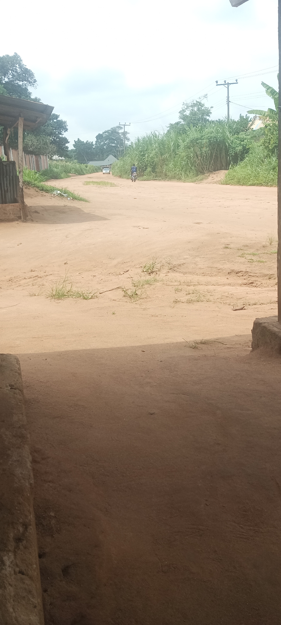 The image depicts a dirt road in Amauzu, Nigeria. A person on a motorcycle is traveling away from the camera, and a car is further down the road in the distance. Lush vegetation lines both sides of the road. Electrical poles and wires are visible. The foreground shows a portion of a building or structure, suggesting the viewpoint is from within or adjacent to a building. The overall scene is rural or peri-urban, evidenced by the unpaved road and surrounding vegetation.