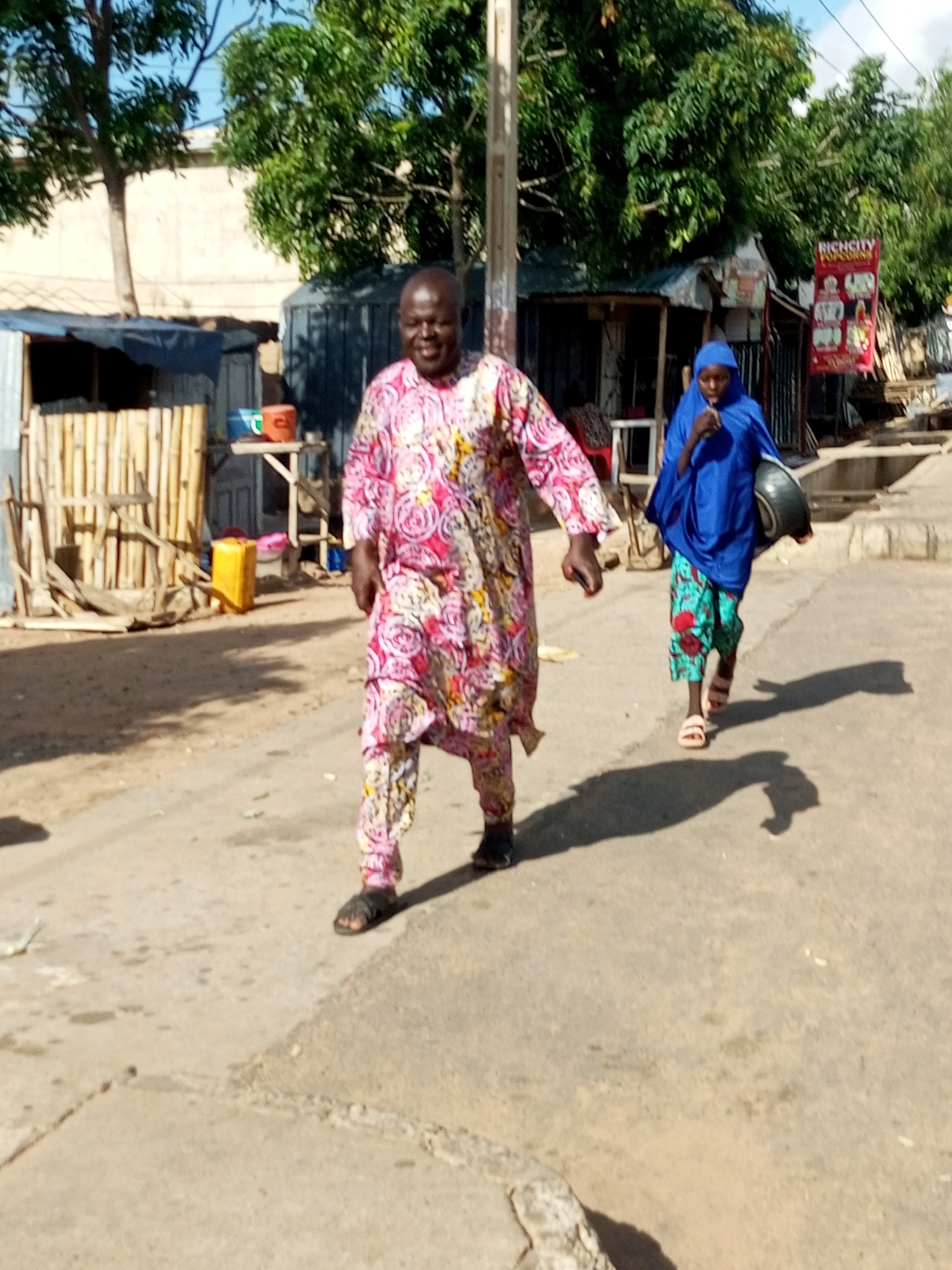 The image depicts a street scene in Tempure, Nigeria, under a bright sky. Two individuals are walking on a dusty, paved road. In the foreground, a man is walking towards the viewer's left, wearing a colorful pink and white patterned garment and dark sandals. He has dark skin and is smiling. Behind him and to his right, a person wearing a blue headscarf and a blue outer garment is walking away from the viewer, carrying a dark basin in their right hand and wearing patterned green and red trousers and light-colored sandals. The background reveals makeshift structures, including stalls made of corrugated metal and bamboo, typical of a market or informal settlement. Trees with lush green foliage are visible, and a white building with a large advertisement is partially visible to the right. Shadows are cast on the ground, indicating the sun is high.