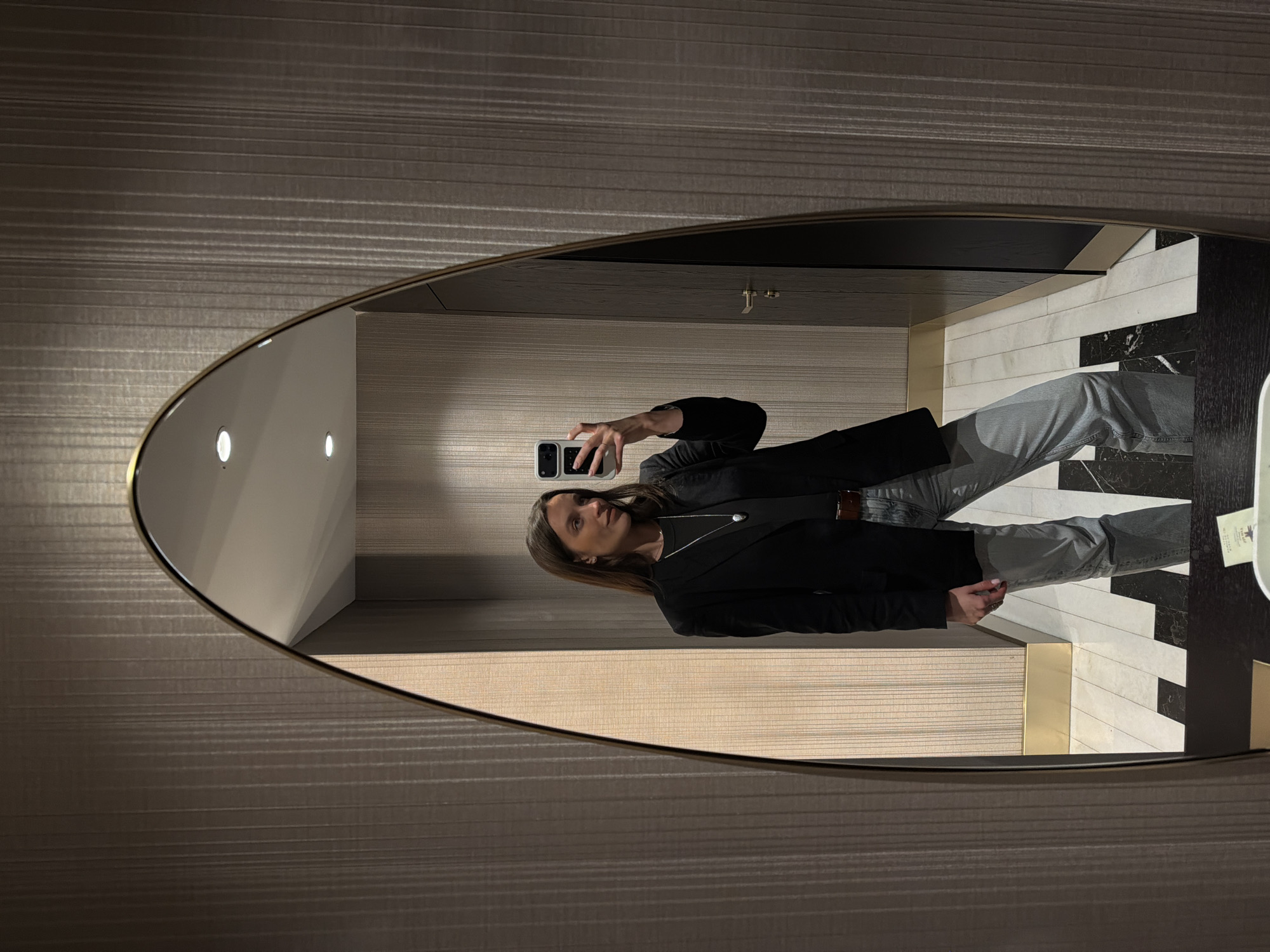 A woman is taking a selfie in an oval mirror. The mirror is reflecting her standing in a room with horizontally textured walls. She is wearing a black blazer, a necklace, and light gray jeans. The floor is tiled in a black and white geometric pattern. The room appears to be in a hotel. While it is not possible to confirm the location is in Budva, Montenegro, without additional information, the style of the room is consistent with modern hotels that might be found there.