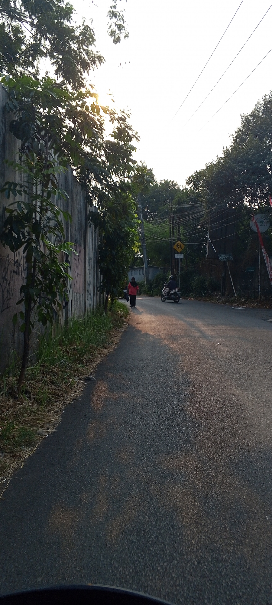 The image depicts a street scene in Depok, Indonesia, during what appears to be late afternoon or early morning, given the low angle of the sun. The sun is casting long shadows and creating a golden glow on the asphalt road.
On the left side of the road, there is a tall, weathered concrete wall covered in graffiti. Adjacent to the wall is a patch of overgrown dry grass and some smaller trees.
On the right side of the road, there is a paved asphalt street with some debris scattered on it. Two people are visible on the street. One person, wearing a red hijab and a pink top, is walking away from the camera on the left side of the road. Further down the road, a person is riding a motorcycle in the same direction.
The background is filled with lush green trees and foliage. Utility poles with a complex network of wires are also prominent in the background, along with a yellow traffic sign featuring two figures, likely indicating a pedestrian crossing or school zone. In the far distance, a portion of a building with a blue roof is visible. The sky is bright and appears overcast in some areas, with the sun peeking through the trees.