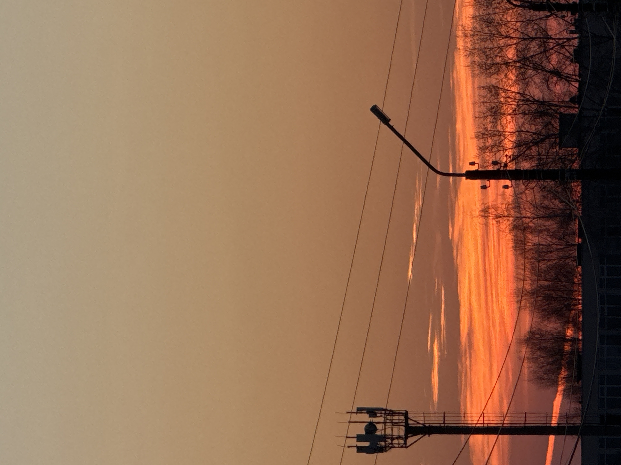 The image depicts an orange sky, likely at sunset or sunrise, in Прикамский, Russia. Silhouetted against the sky are bare trees, utility poles, and a street light. There's a building visible on the right. The scene is captured from a static, ground-level perspective.