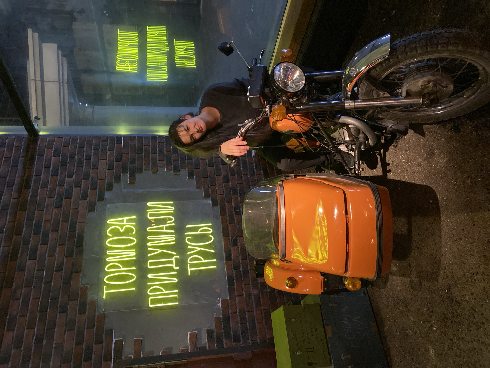 The image depicts a woman sitting on an orange motorcycle with a sidecar, likely in Moscow, Russia, as suggested by the Cyrillic text. The motorcycle is parked on what appears to be a city street with brick walls. There is a neon sign on the wall that reads "ТОРМОЗА ПРИДУМАЛИ ТРУСЫ" in Cyrillic, which translates to "Brakes were invented by cowards." Above, a window with reflection displays more neon writing “ACOMPOT ШЬХОРІ”. A few green military boxes with Cyrillic writing are near the sidecar.