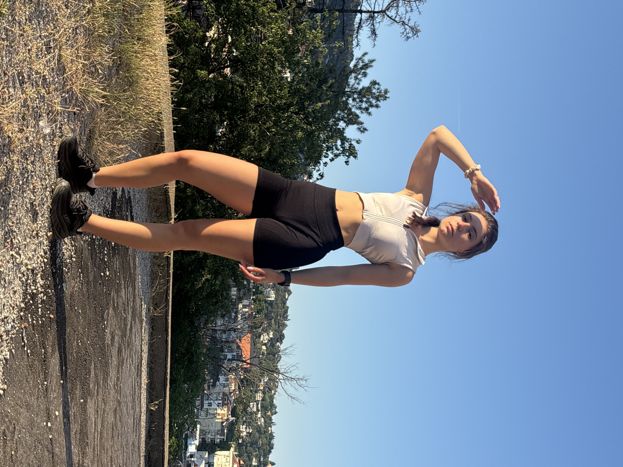 This is a full-body, portrait-oriented, outdoor photograph of a young woman posing on a paved surface with a scenic overlook of a city and the sea. The woman is standing with her left leg slightly bent and her right leg extended. She is wearing black athletic shorts, a light-colored sports bra, and black sneakers. Her left arm is raised and bent at the elbow, with her hand resting on her forehead, as if shielding her eyes from the sun. Her right arm is down by her side. She has dark hair pulled back and is looking towards the camera with a neutral expression. A thin bracelet is visible on her right wrist.
The setting appears to be a park or elevated viewpoint overlooking a coastal city. In the background, there is a dense cluster of trees to the left and a clear, bright blue sky with a faint contrail across the upper portion. Below the trees, a dense residential area with buildings of various colors, predominantly white and orange roofs, cascades down a hillside towards the sea.
The lighting suggests it is daytime, likely late morning or early afternoon, given the bright sun and the shadows cast. The weather is clear and sunny. The overall impression is one of a casual, active moment captured during a pleasant day outdoors in Varna, Bulgaria. No visible text is present in the image.