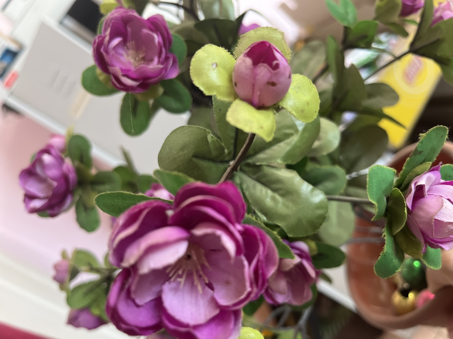 A close-up image depicts an artificial flowering plant in an indoor setting in Kyez, Russia. The plant, positioned centrally, features multiple clusters of small, fabric-based flowers. The dominant flower color is a deep magenta or purple, with some showing lighter pink hues, particularly towards their centers, and visible yellow stamens. Several flower buds are present, displaying lighter pink petals encased by yellowish-green sepals. The foliage consists of numerous small, oval, dark green fabric leaves. The plant is contained within a visible section of a terracotta-colored pot, located in the lower left of the frame. The background is heavily blurred, revealing indistinct light-colored surfaces, potentially walls or furniture, and vague shapes, including a yellow object on the lower right and several light-colored rectangular objects towards the upper right. No individuals are present, and no discernable actions or interactions are occurring.