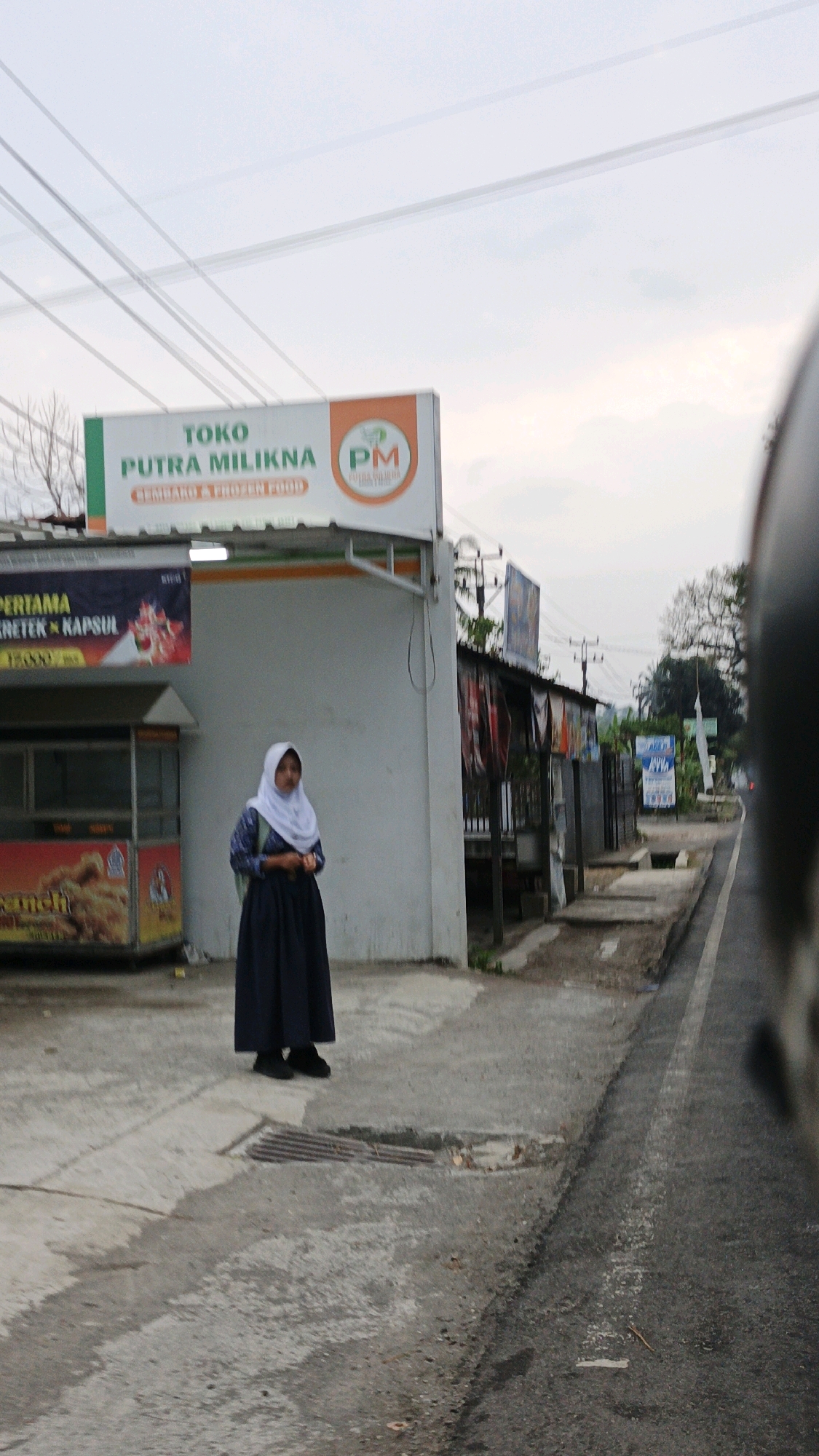 A medium shot captured at street level shows a young woman standing on a sidewalk in front of a storefront. The woman is wearing a white hijab and a dark blue long skirt and blouse, with a dark backpack on her back. She is facing the camera with her head slightly tilted. Behind her, a white building with an orange trim is visible, with a sign that reads "TOKO PUTRA MILIKNA" in green lettering. To the left of the building, another sign advertises "PERTAMA KRETEK & KAPSUL" with an image of a cigarette pack.
The sidewalk is paved with concrete and shows some wear and tear, with a metal grate in the foreground. The street, which is paved with asphalt, runs alongside the sidewalk and appears to be wet, suggesting recent rain. In the background, other buildings and trees are visible, along with power lines stretching across the sky. The overall scene suggests a quiet street in a town or city, possibly during the early morning or late afternoon given the soft lighting. The location is identified as Cibenda, Indonesia.