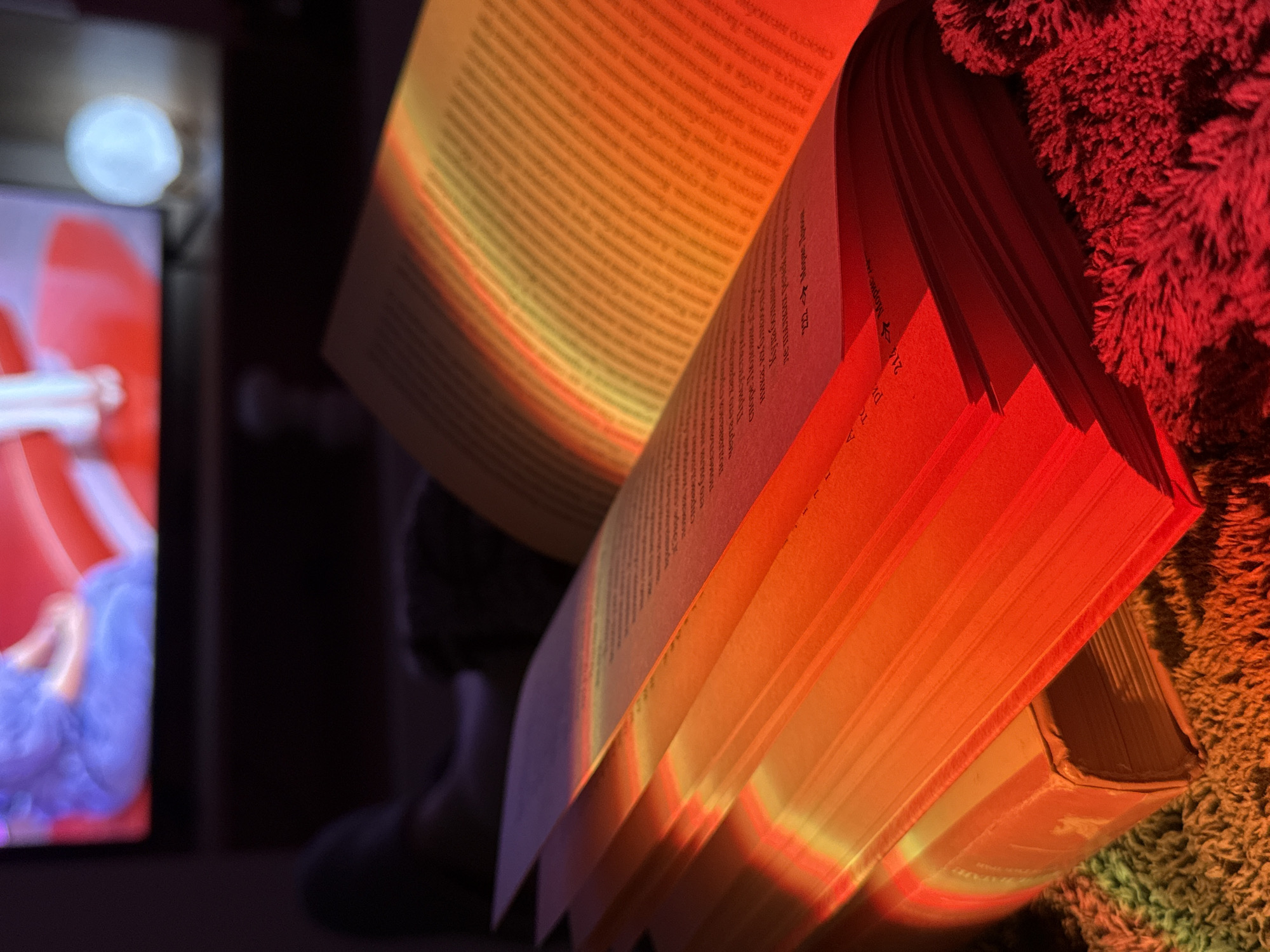 An open book is illuminated with red and orange light. The pages are visible, filled with text in an unidentified language. The book sits on a textured surface, possibly a rug or blanket. In the background, there is a partially visible screen displaying a blurry image with red and blue hues. The setting appears to be indoors, though the exact location is indiscernible. There are no people visible in the foreground.