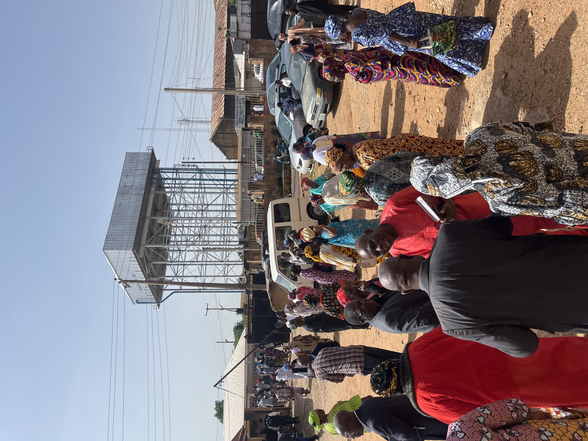 In Bukuru, Nigeria, a group of people are gathered outdoors on a dusty, reddish-brown ground. A large, elevated metal water tank dominates the background, supported by a complex metal frame. Several buildings with corrugated metal roofs are visible behind the people and the water tank. Power lines crisscross the sky.
The crowd consists of both men and women, many of whom are dressed in brightly colored, patterned clothing, including headwraps and flowing garments. Some individuals are engaged in conversation, while others appear to be observing or waiting. A white van is parked among the people, and several cars are visible in the background. One building has a sign that reads "SUA FURNITURE WORKS". The overall atmosphere suggests a public gathering or event.