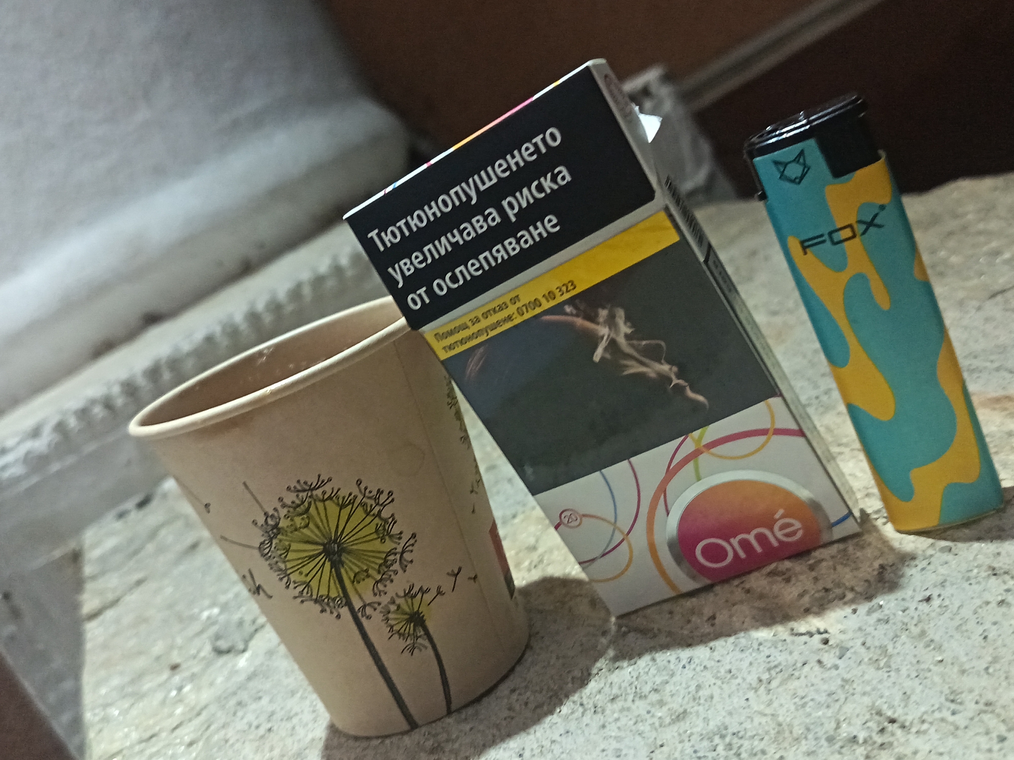 The image displays a still life composition featuring a paper cup, a cigarette pack, and a lighter, resting on a textured, light-colored surface, possibly concrete or stone, in Градина, Bulgaria.
The paper cup, positioned on the left, is light brown and decorated with a graphic illustration of dandelions. In the center, a pack of Omé cigarettes stands upright. The top portion of the cigarette pack features a prominent black health warning in Bulgarian, which reads: "Тютюнопушенето увеличава риска от ослепяване" (Smoking increases the risk of blindness). Below this, a yellow strip contains the text: "Помощ за отказ от тютюнопушене: 0700 10 323" (Help to quit smoking: 0700 10 323). The lower part of the pack shows the brand name "Omé" and the number "20". To the right of the cigarette pack is a lighter, emblazoned with the brand "FOX" and a fox head logo. The lighter has a distinctive design featuring teal and yellow abstract or camouflage patterns.
No people are present, and no specific activity is taking place; the scene is a static arrangement of objects. The objects cast shadows, indicating natural light, possibly daytime in a shaded outdoor setting. The background is out of focus, providing little additional detail about the immediate surroundings.
