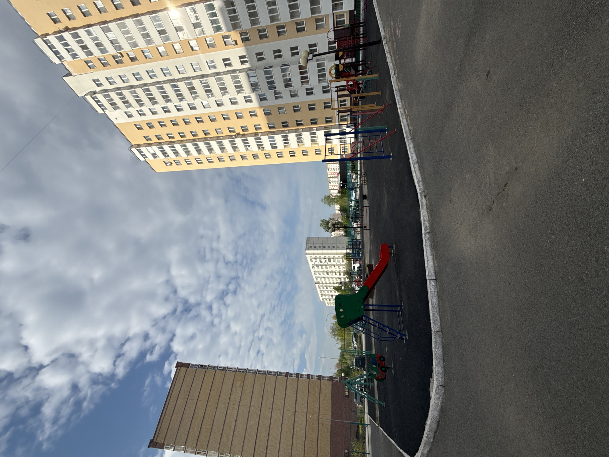 The image captures an outdoor playground in Tobolsk, Russia. Tall residential buildings flank the playground. The sky is partly cloudy. Playground equipment includes a slide, climbing structures, and swings. The ground is paved with dark asphalt. No people are visible in the frame.