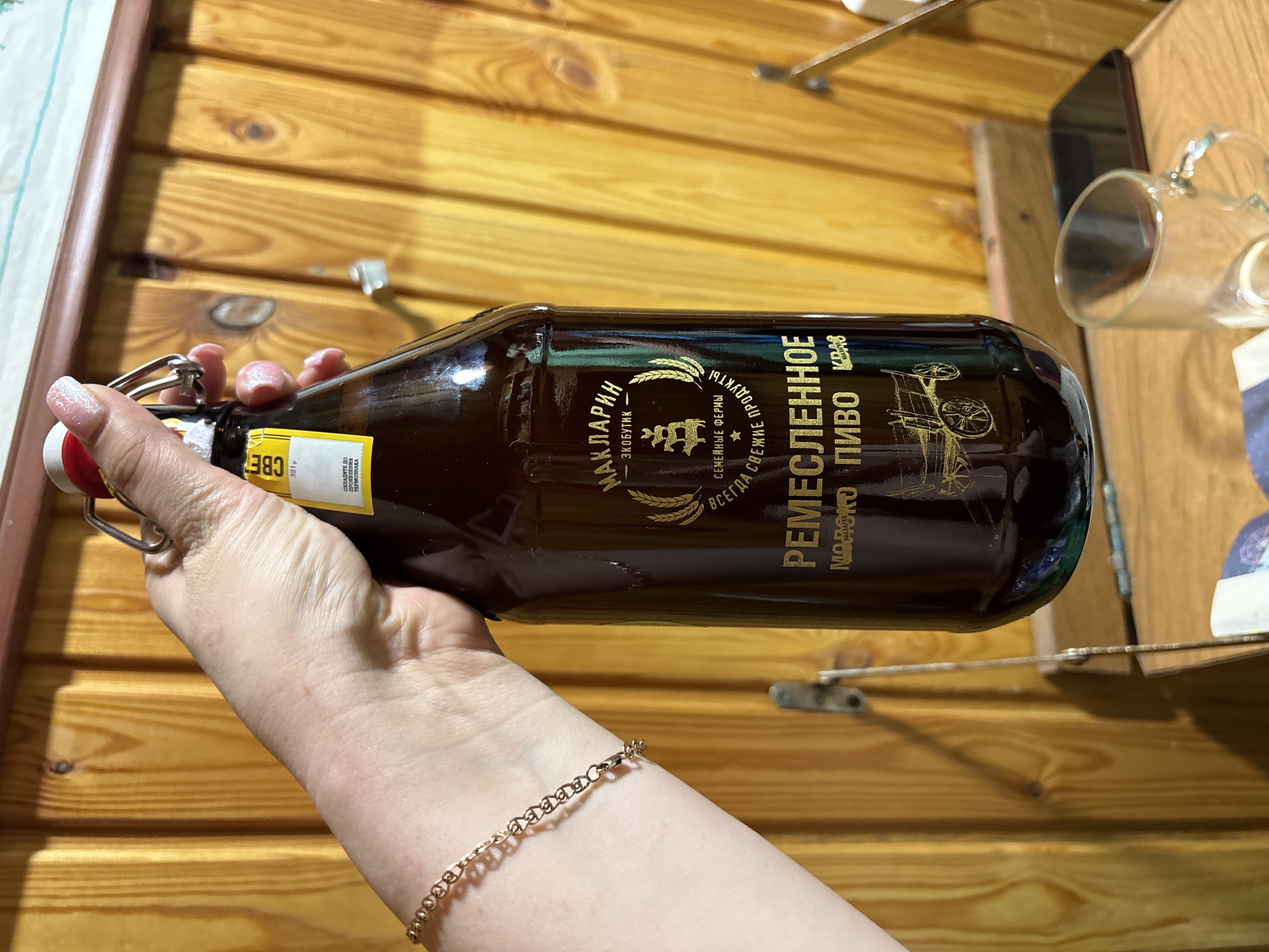 A hand holds a brown, glass bottle of "Remeslennoe" beer, with Russian text on the label, against a background of wooden planks. The text on the label includes "Maklarin" and "Eko-boutique," suggesting a brand name and environmentally-friendly positioning. A gold-colored bracelet is visible on the wrist. There is a shelf with a drinking glass mounted to the wall in the background. Based on the label in the photo, this image may have been taken in Dubovaya, Russia.