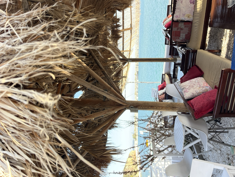 The image shows an outdoor seating area located in Limenaria, Greece, at the coordinates 40.626, 24.576. The area is covered by a large, thatched-roof umbrella, providing shade from the sun. There are white chairs and a small white table in the foreground, and a larger wooden couch and table in the background. The couch is covered with cushions, some of which have floral patterns. The wooden structure of the umbrella extends to the back of the couch, creating an enclosure. The scene is overlooking the azure waters of the Aegean Sea, with a shoreline of a small town in the distance. The environment is relaxed and inviting, with a pleasant Mediterranean breeze blowing through the area.