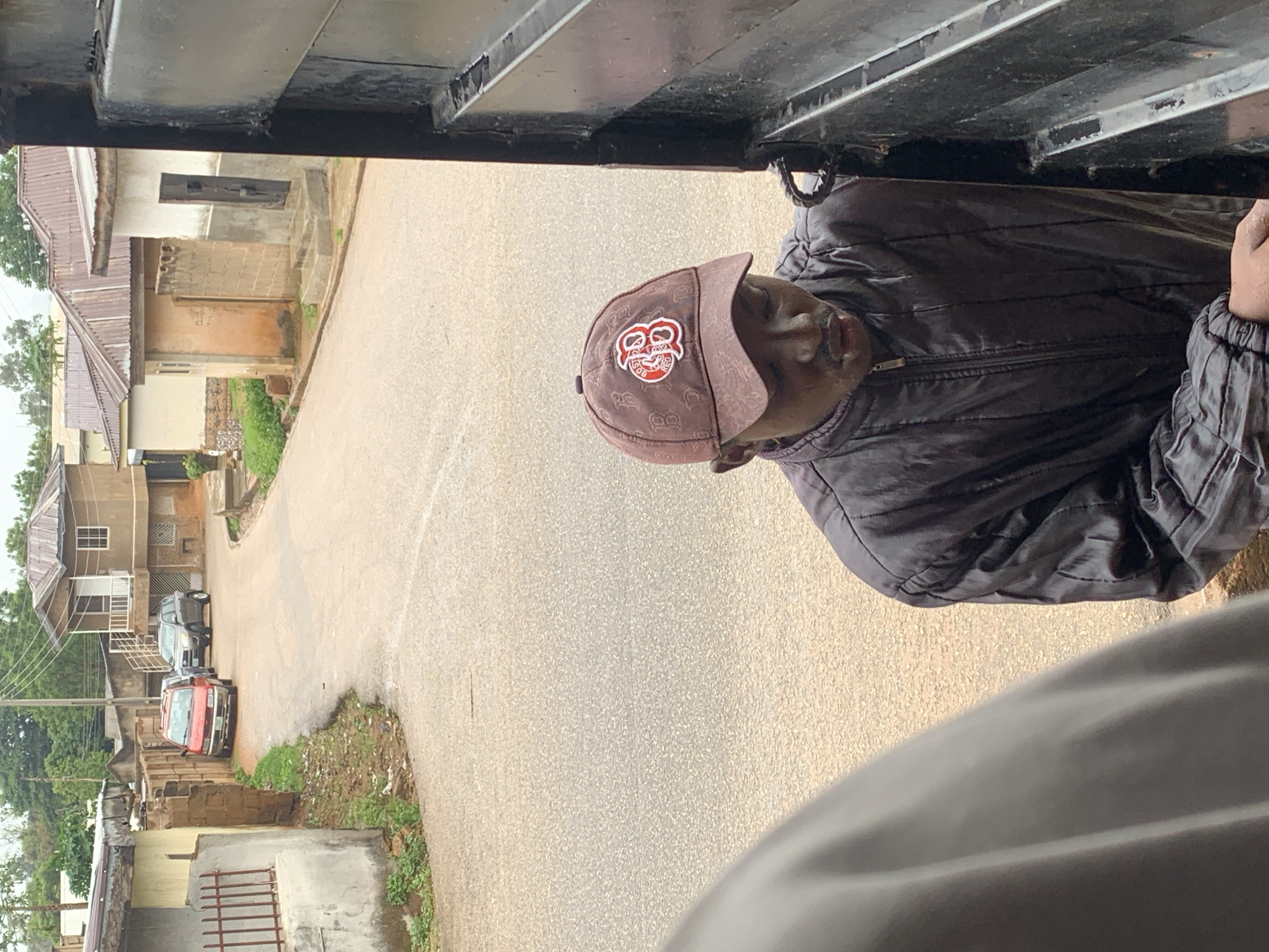 The image shows a man wearing a dark jacket and a patterned baseball cap with a red and white logo. He is standing next to a black metal gate, appearing to be opening or closing it. The background depicts a street in a residential area, with houses and parked vehicles visible. The street is paved with asphalt and has patches of grass and dirt along the sides. The architecture of the houses suggests a location in Nigeria, potentially in the city of Jos, characterized by their single-story design with corrugated metal roofs and concrete construction. The overall scene is that of an everyday street view in a Nigerian city.