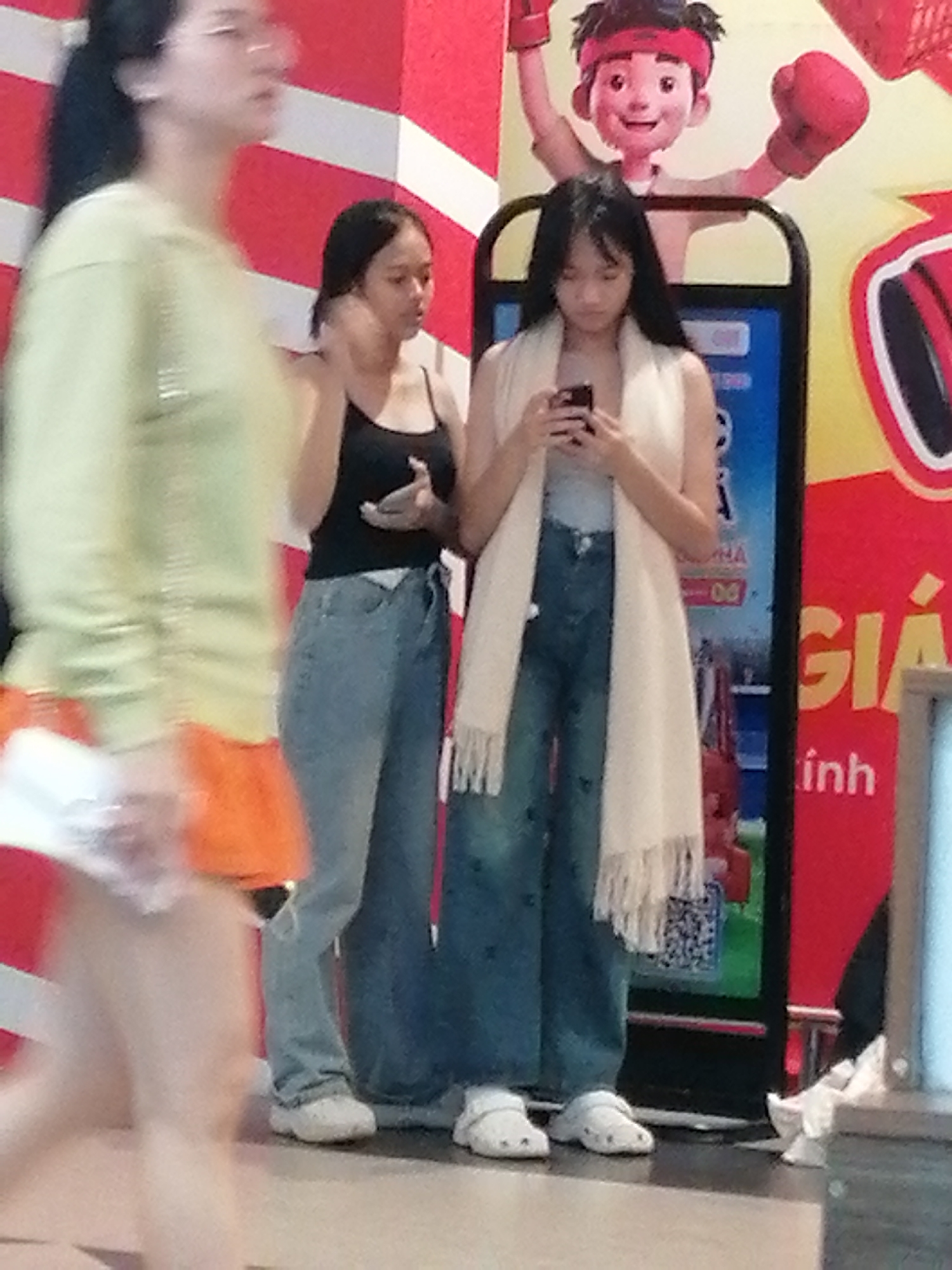 The image captures a candid street scene in Da Lat, Vietnam, focusing on three young women. In the foreground and to the left, a woman with dark hair and glasses is partially visible, her body angled away from the camera, wearing a pale green long-sleeved top and an orange skirt. Her movement is blurred, suggesting motion.
To the right of the first woman, two other young women stand together. The one on the left, with dark hair tied back, wears a black tank top and light blue jeans. She is looking downwards and holding something in her hands. The woman on the right, also with dark hair, is wearing a light-colored scarf over a grey top and dark blue jeans. She is looking down at her phone, which she holds in her hands.
Behind the two women, a colorful backdrop features a cartoonish character with a red headband and boxing gloves, suggesting an entertainment or advertisement display. Text in Vietnamese, including "GIÁ" and "inh," is visible on a red sign to the right of the women, indicating the location may be a commercial or public space within Da Lat. The ground appears to be tiled. The overall lighting suggests an indoor or shaded outdoor environment.