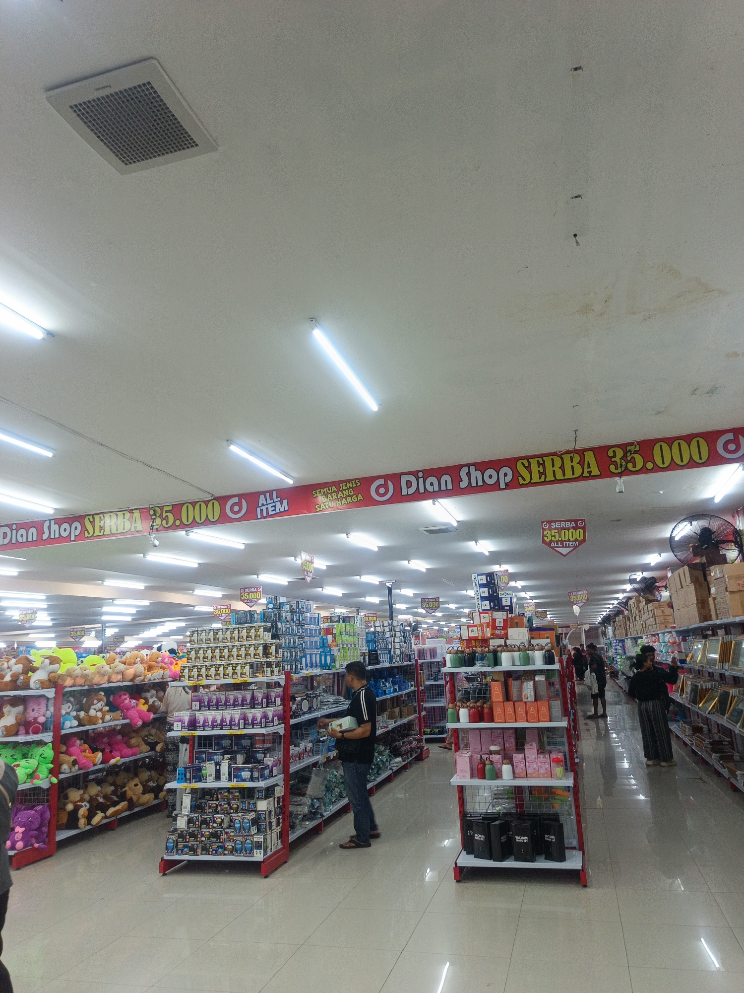 The image depicts the interior of a retail store, likely a department store or variety shop, in Limboto, Indonesia. The store features brightly lit aisles with shelves stocked with a wide variety of merchandise, including stuffed animals, home goods, and electronics. A prominent red banner stretches across the ceiling, advertising "Dian Shop SERBA 35.000 ALL ITEM," indicating a fixed price of 35,000 Indonesian Rupiah for all items. Several customers are visible browsing the aisles. One man in a black shirt and blue jeans is examining a product, while a woman in a dark outfit is further down the aisle, appearing to shop. The environment is clean and organized, with reflective tiled floors. The ceiling has a ventilation grate and fluorescent lighting.