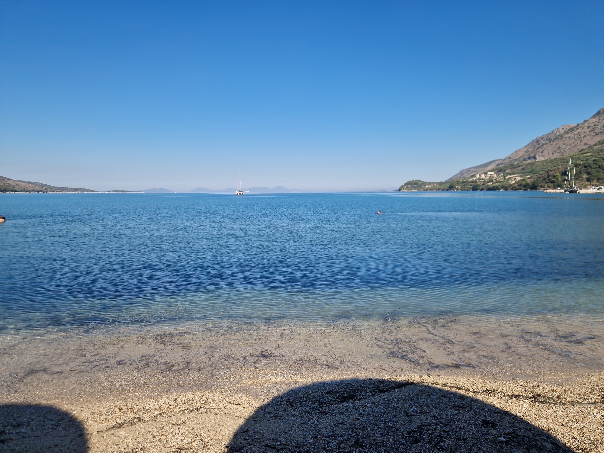 A wide-angle, eye-level, outdoor shot captures a serene view of the sea and a beach in Plataria, Greece. The clear blue sky dominates the upper half of the frame. The sea is a calm, deep blue, with gentle ripples reflecting the sunlight. In the distance, a white sailboat is visible, with another boat, possibly a yacht, closer to the right shore. A hilly, tree-covered landscape lines the horizon on the right side of the image, with a small village nestled on the hillside. The foreground shows a beach with small pebbles and sand, partially shaded by a dark, curved shadow. A person is visible swimming in the sea to the left of the frame, and another is swimming further to the right. The water is clear enough to see the seabed near the shore.